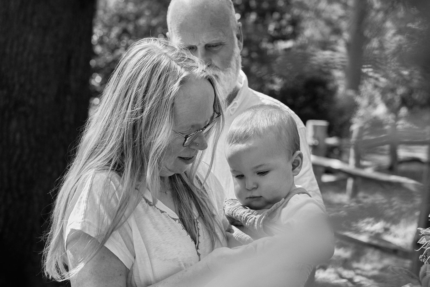 Black and white photo of grandparents lovingly holding a baby outdoors in a wooded setting.