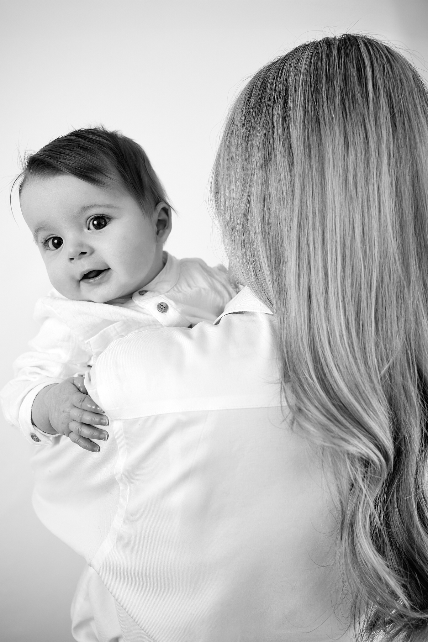 Black and white photo of a blonde mother holding her smiling baby in a white onesie against a light background.