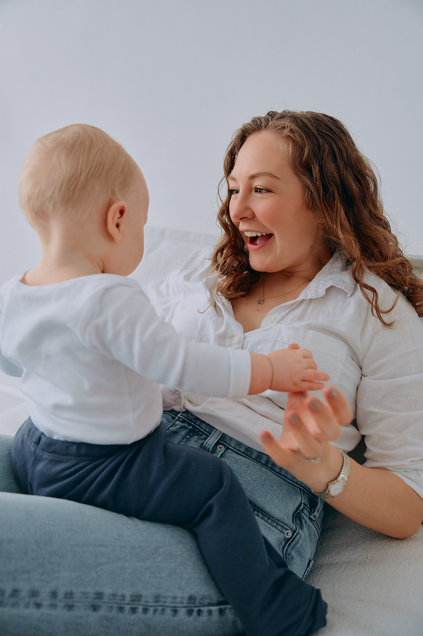 A joyful mother in a white shirt and jeans plays with her baby on a light background.