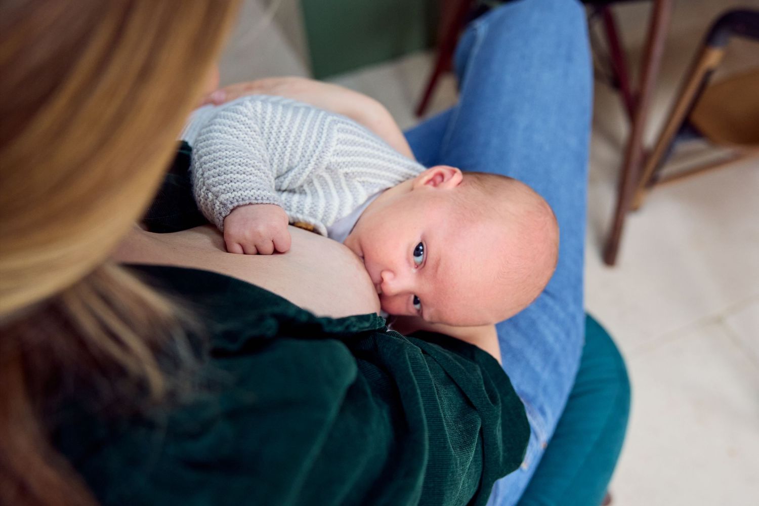 Mother breastfeeding her newborn baby while sitting in a chair, infant wearing striped onesie.