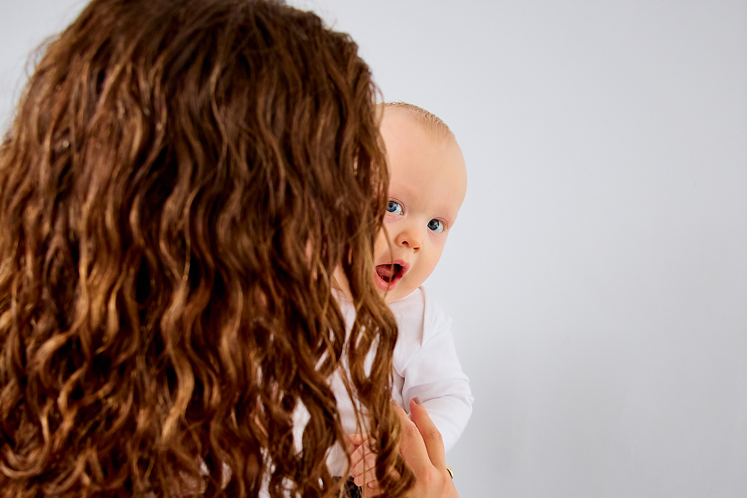 A mother with curly brown hair holds her wide-eyed baby in a white onesie against a light background.