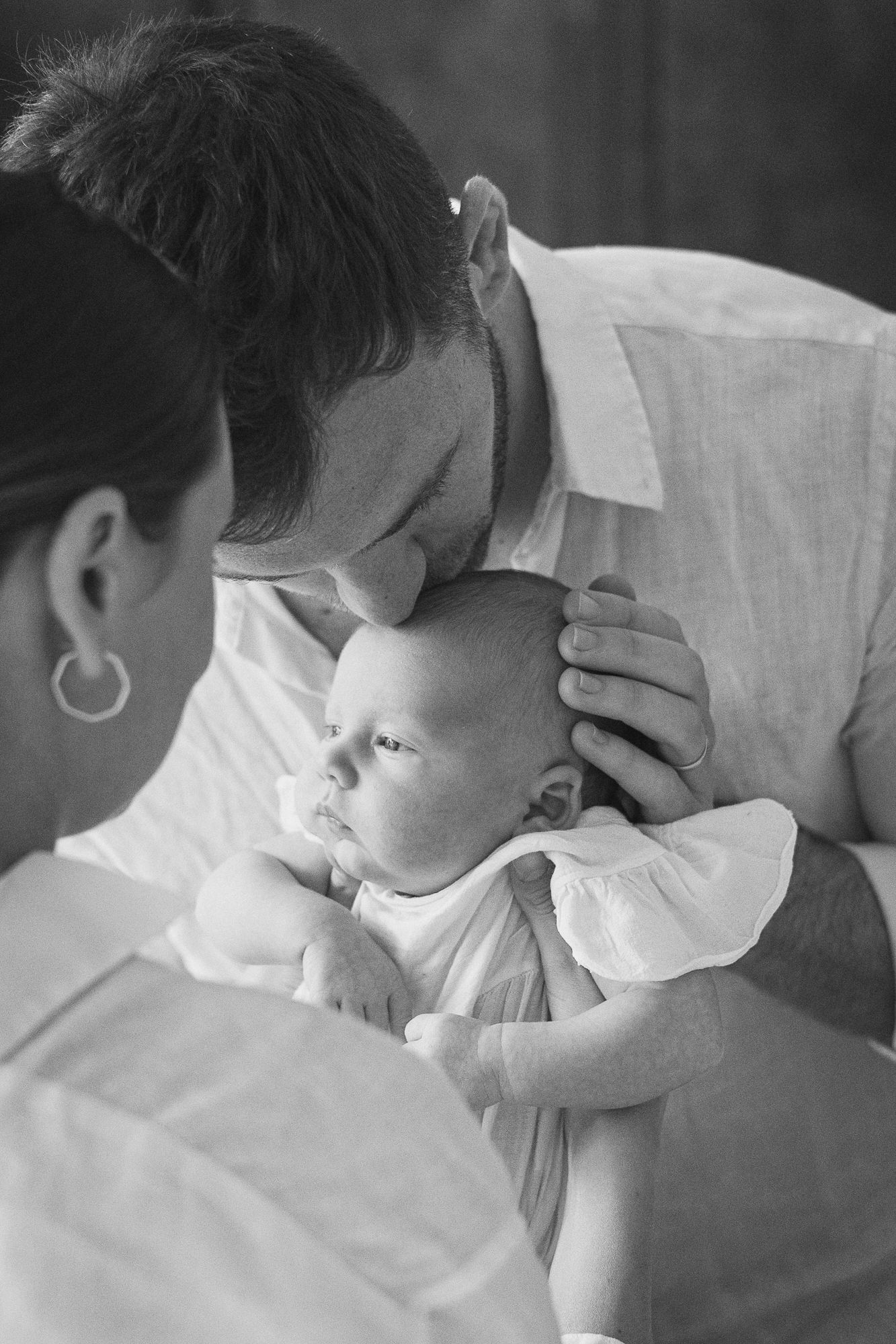 Black and white photo of a father tenderly kissing his newborn baby's head while mother holds the infant.