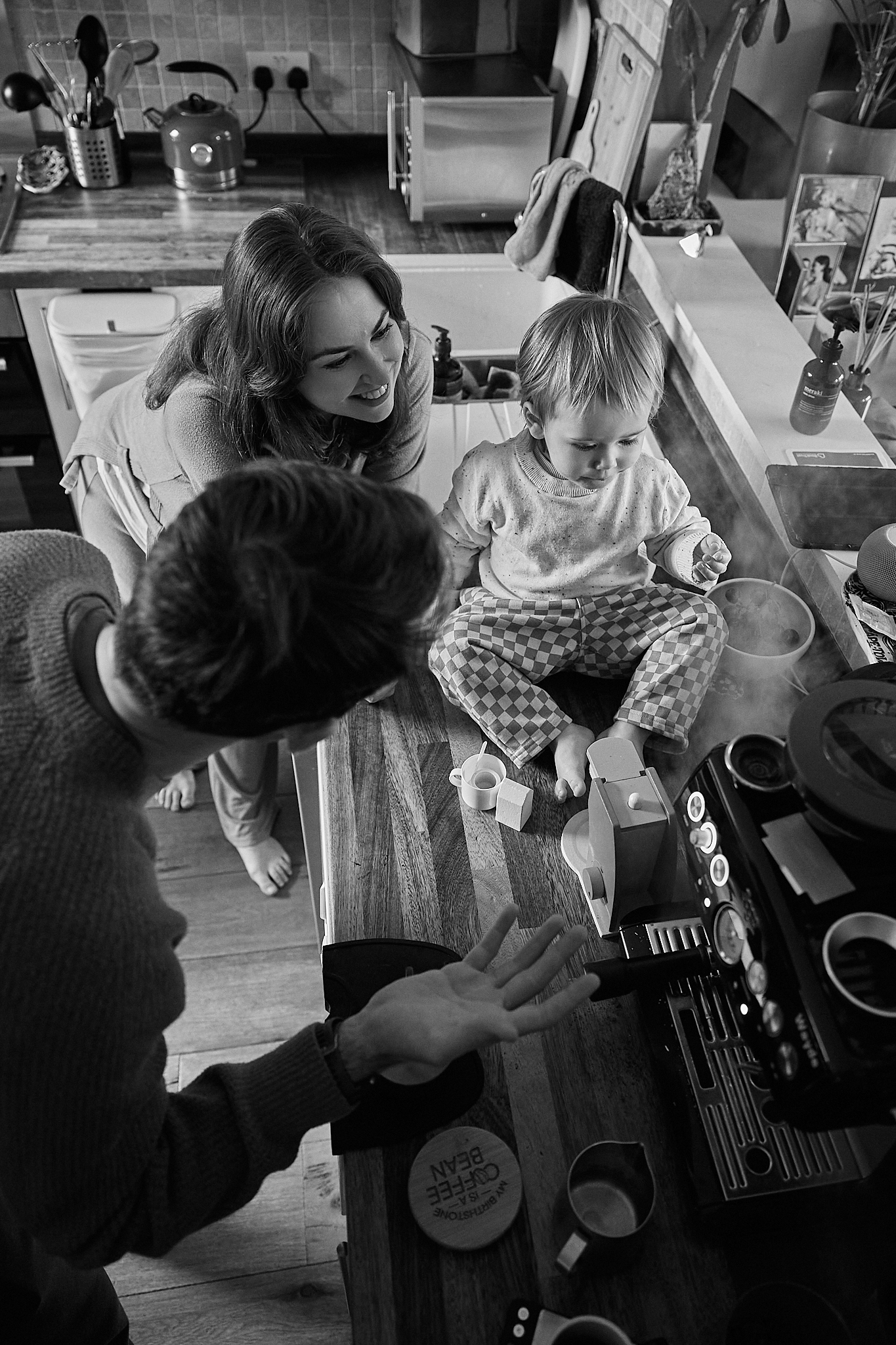 Black and white photo of a family in kitchen, mother smiling at toddler sitting on counter while father reaches out.