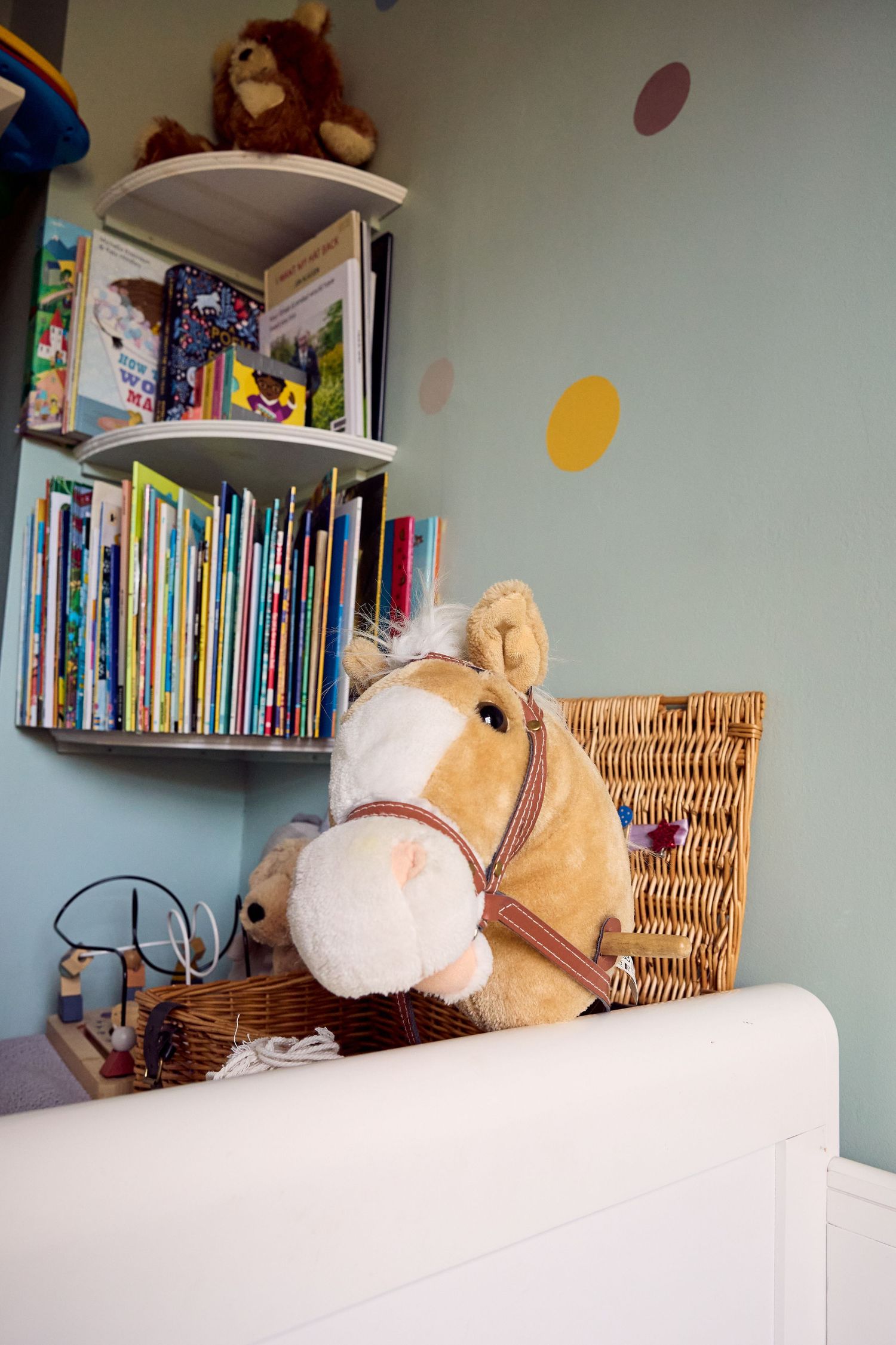Stuffed horse toy on white dresser beside wicker basket and colorful children's bookshelf in pastel nursery room.