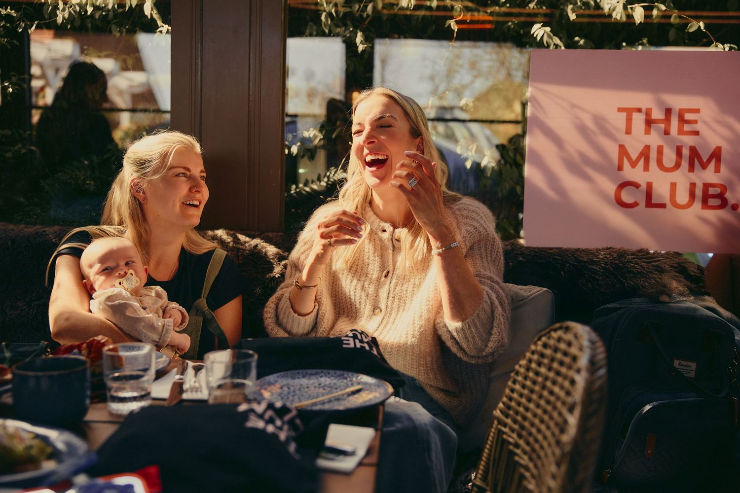 Two women laughing together at The Mum Club outdoor event in warm sunlight.
