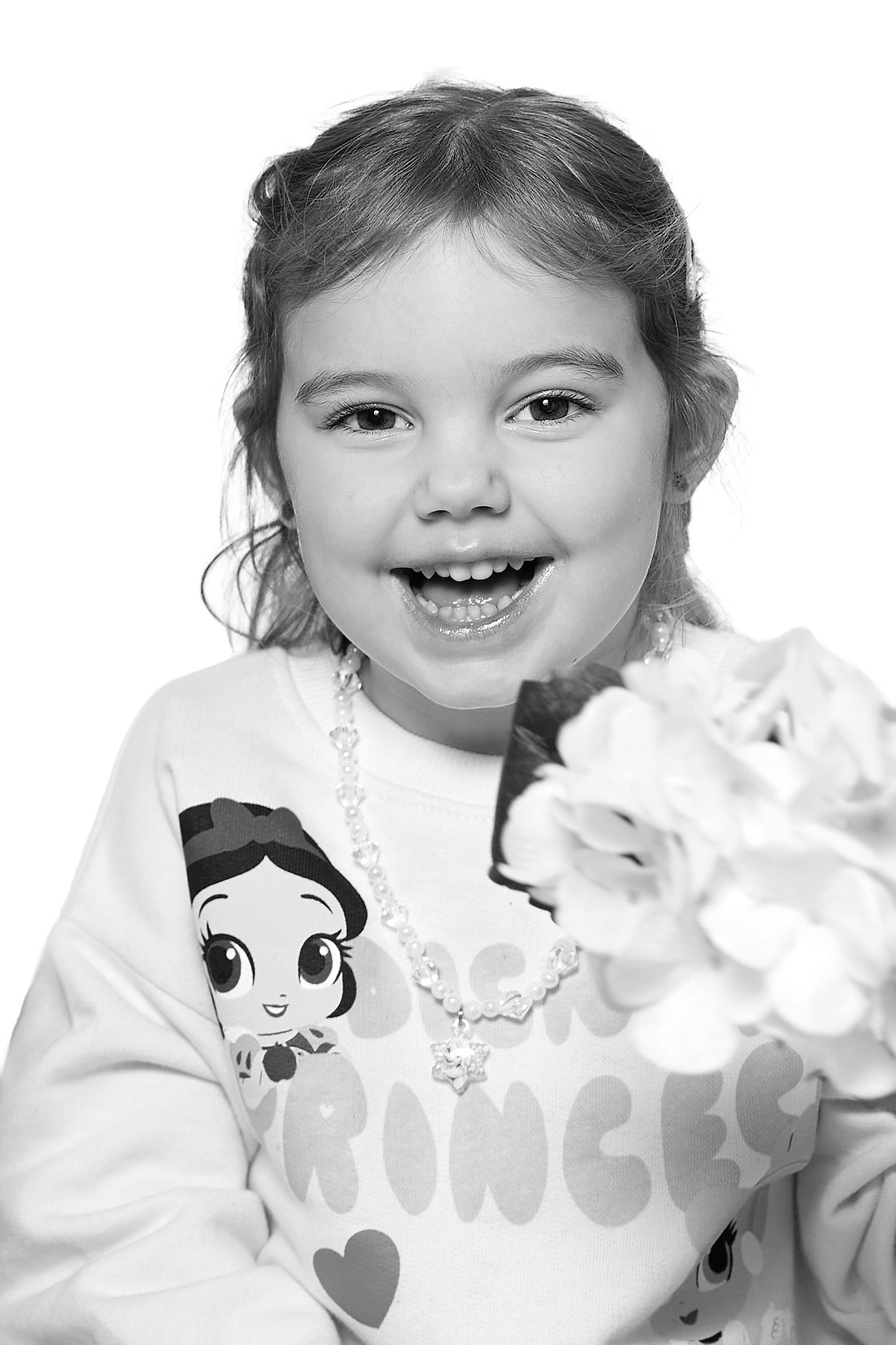 Smiling young girl wearing a Princess shirt and bead necklace holding flowers in a black and white portrait photo.