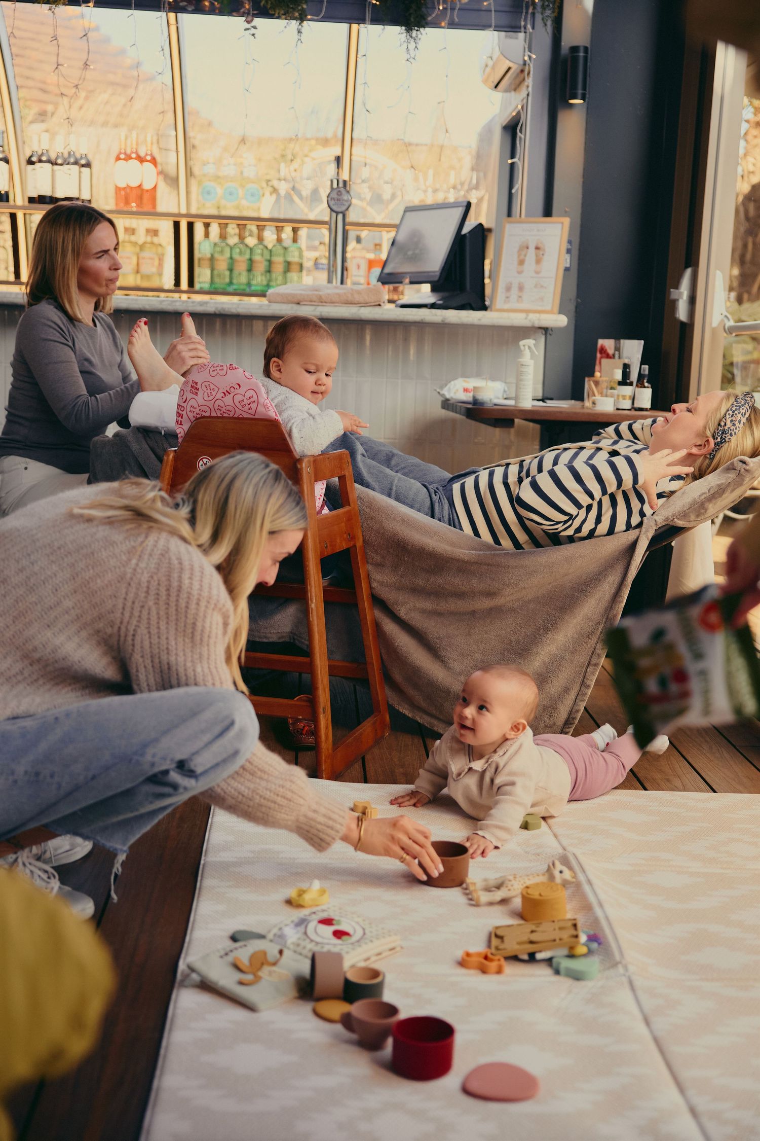 Two mothers play with their babies on a soft mat in a cozy cafe, surrounded by colorful toys.