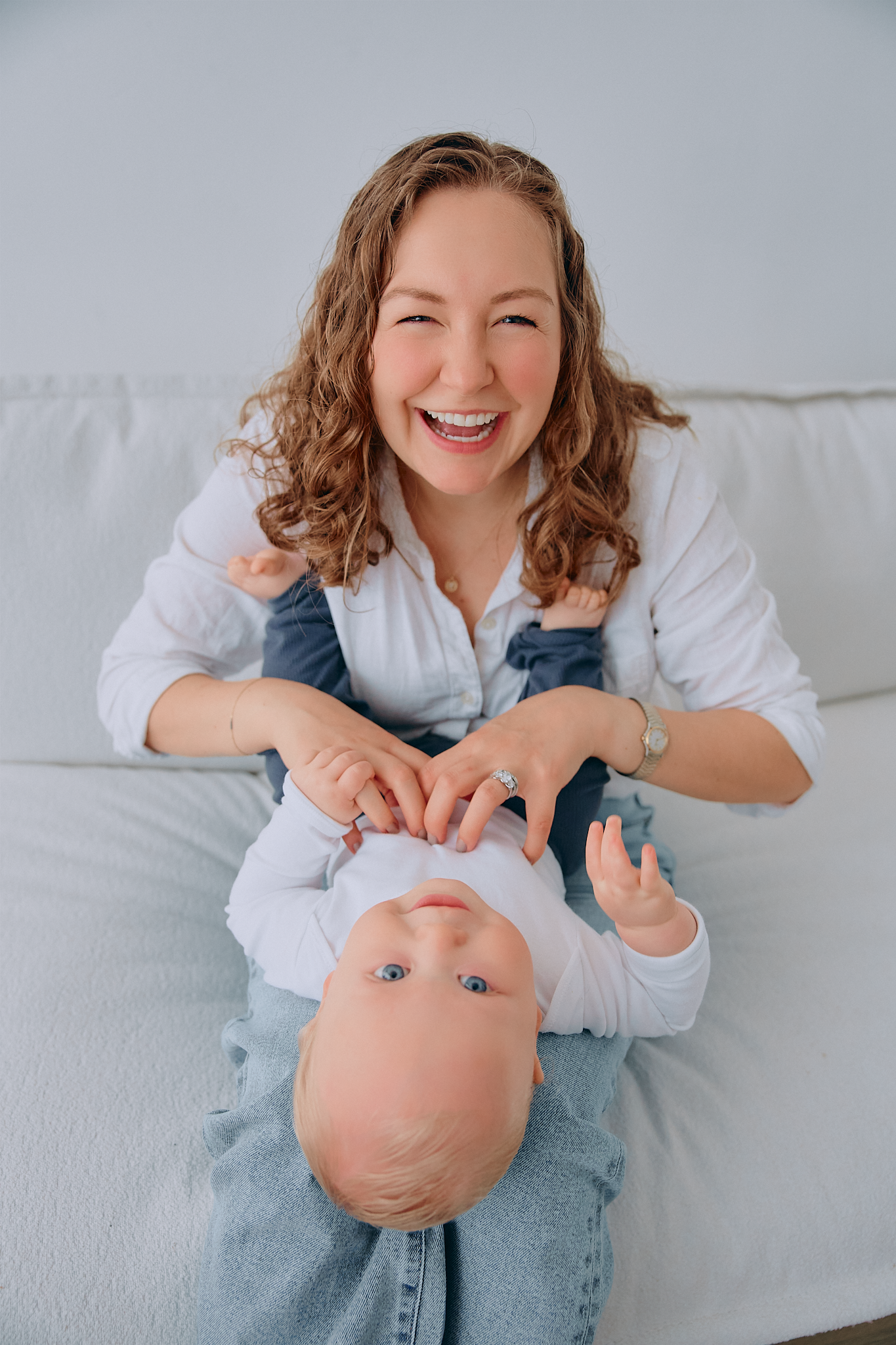 Laughing mother playfully holding her blue-eyed baby upside down on a white sofa.