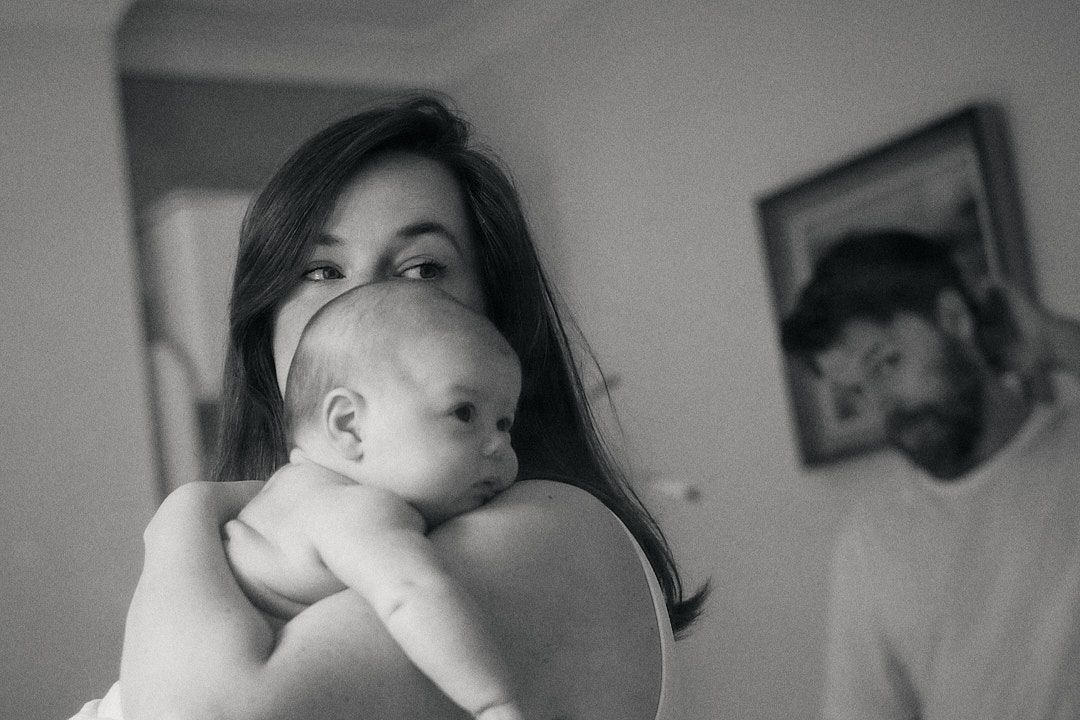 Black and white photo of a young mother holding her newborn baby, with a man visible in the background.