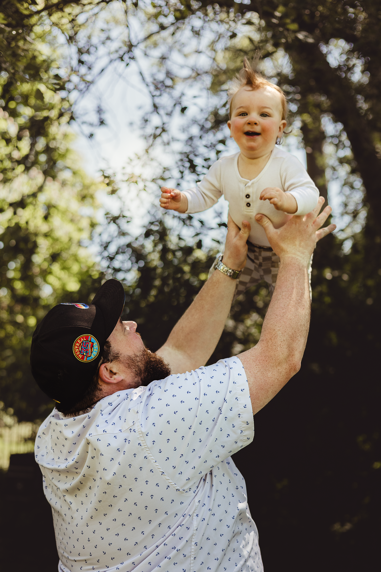 Baby wearing white onesie is lifted joyfully in the air against sunny outdoor backdrop.