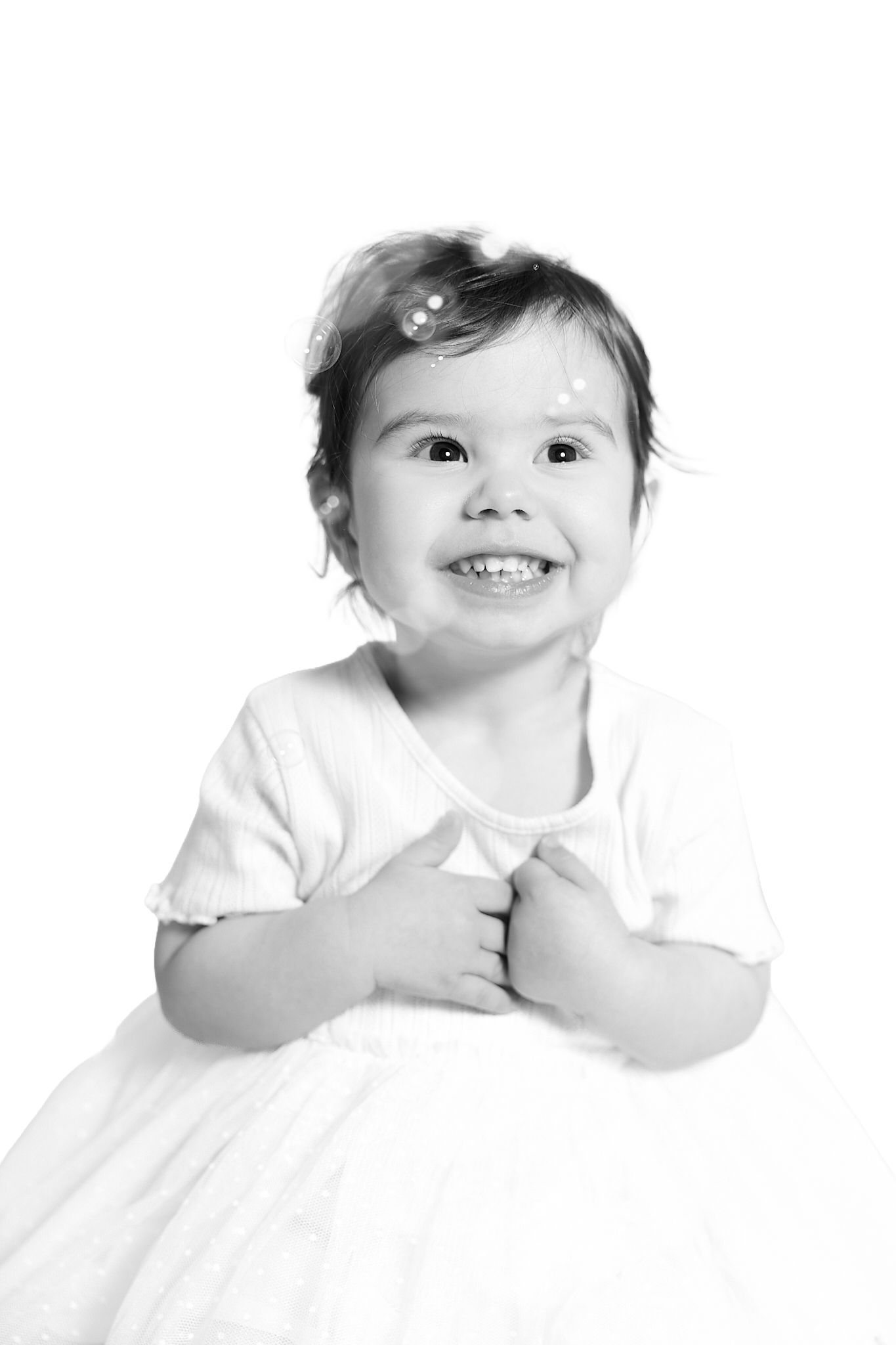 Black and white portrait of a happy smiling toddler girl in a white dress, looking up with joy.