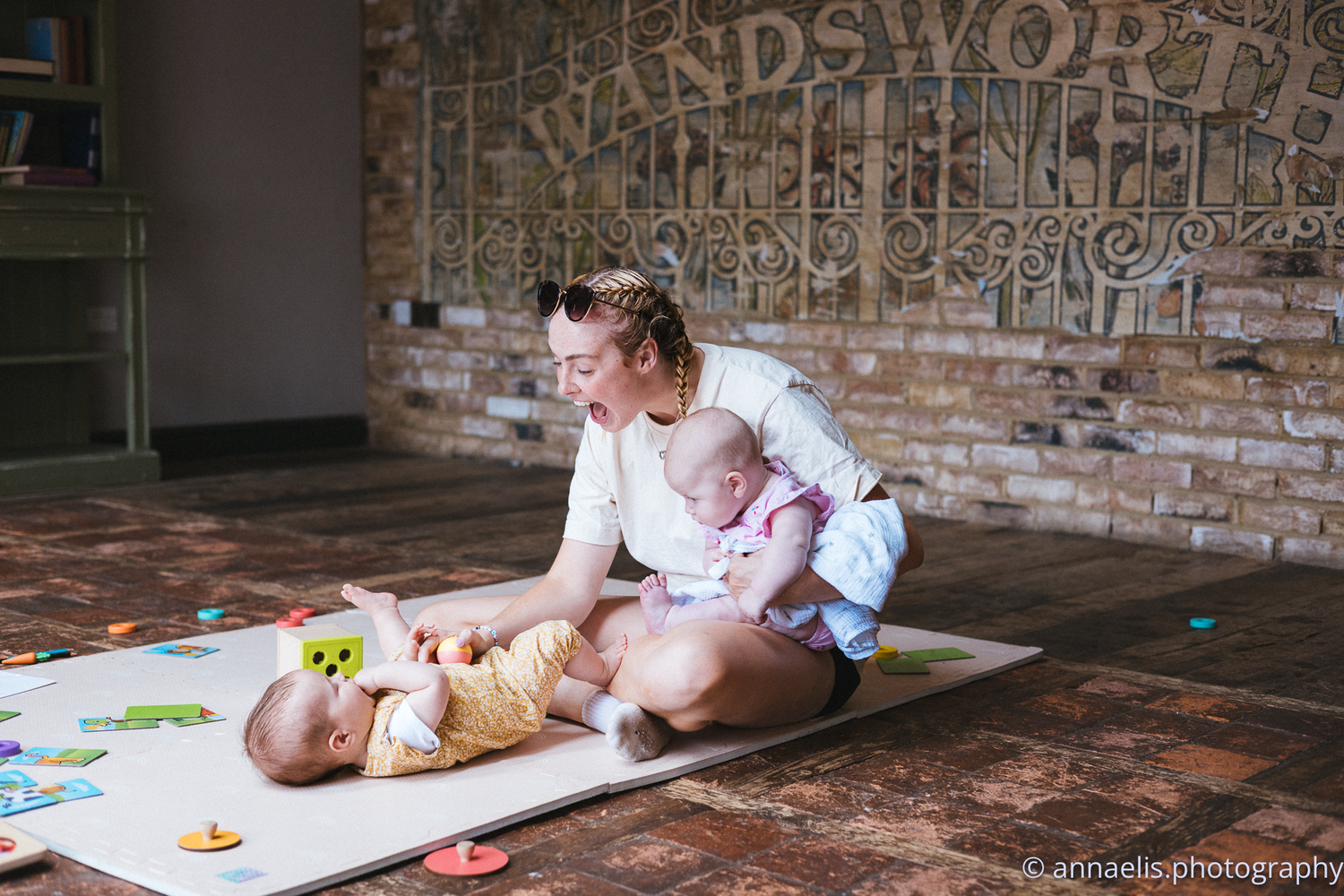 Baby crawling on floor mat with toys and colorful blocks in rustic brick room setting.