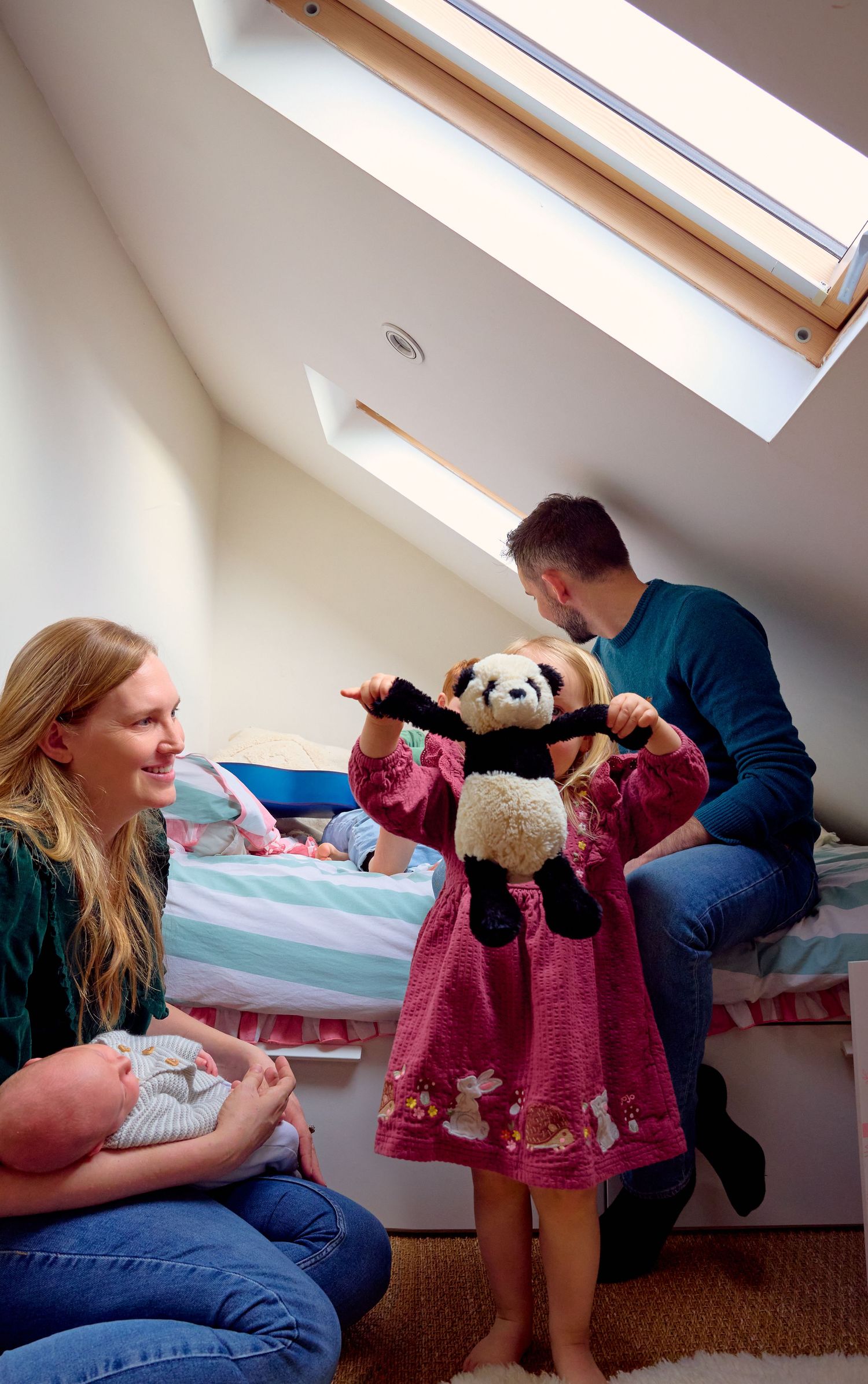 Family with newborn baby and toddler girl holding panda toy in attic bedroom.