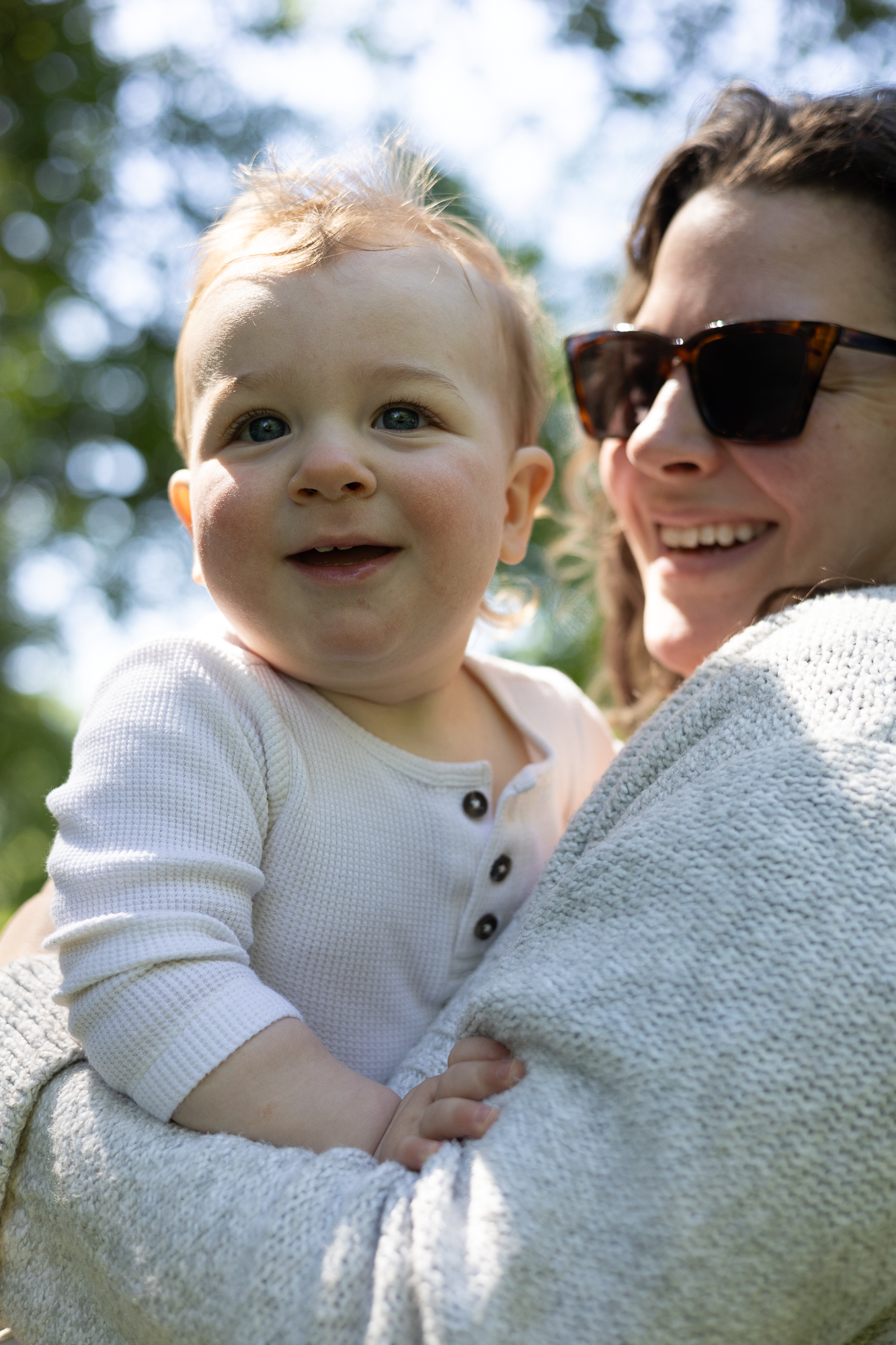 Baby in white sweater smiles while being held against soft gray knit fabric outdoors.