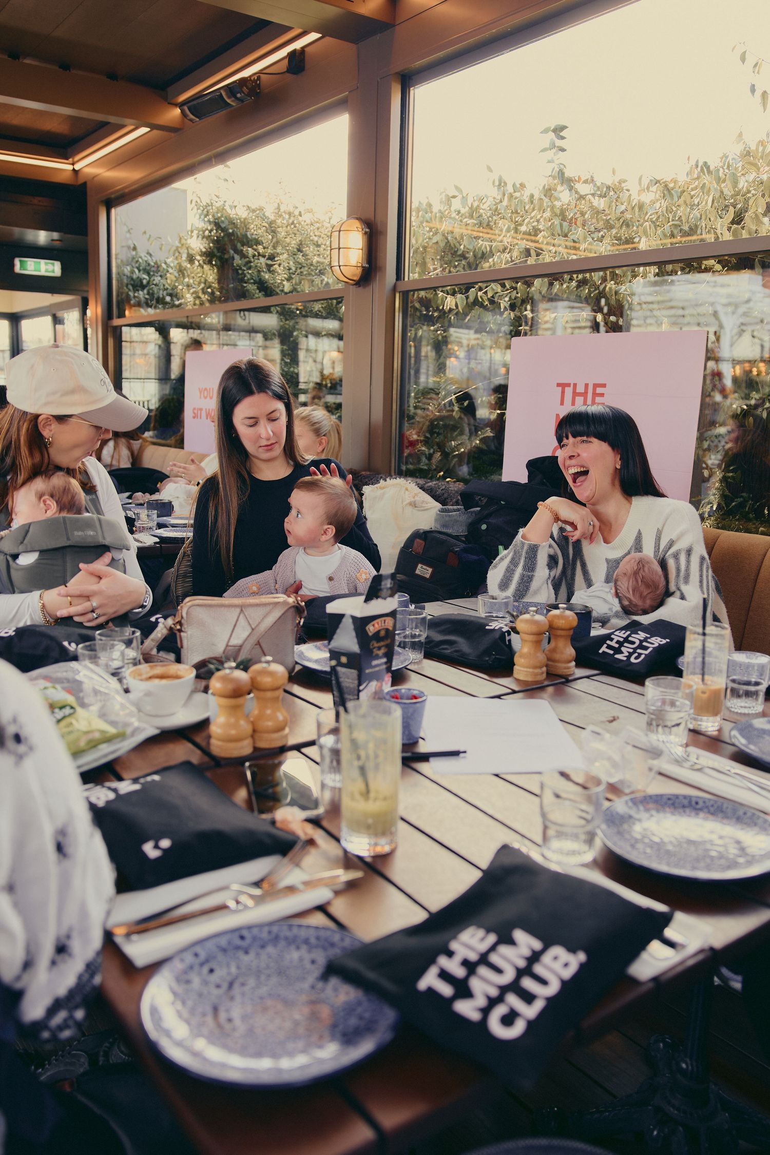 Mothers laughing and bonding at a The Mum Club dining event with babies at a restaurant table.