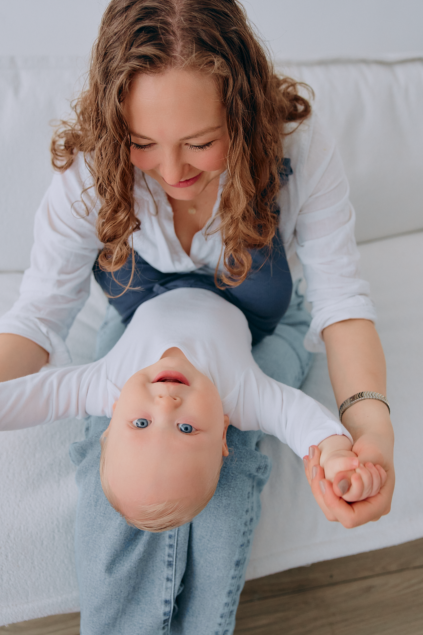 A smiling mother with curly hair holds her blue-eyed baby upside down playfully while sitting on a white couch.