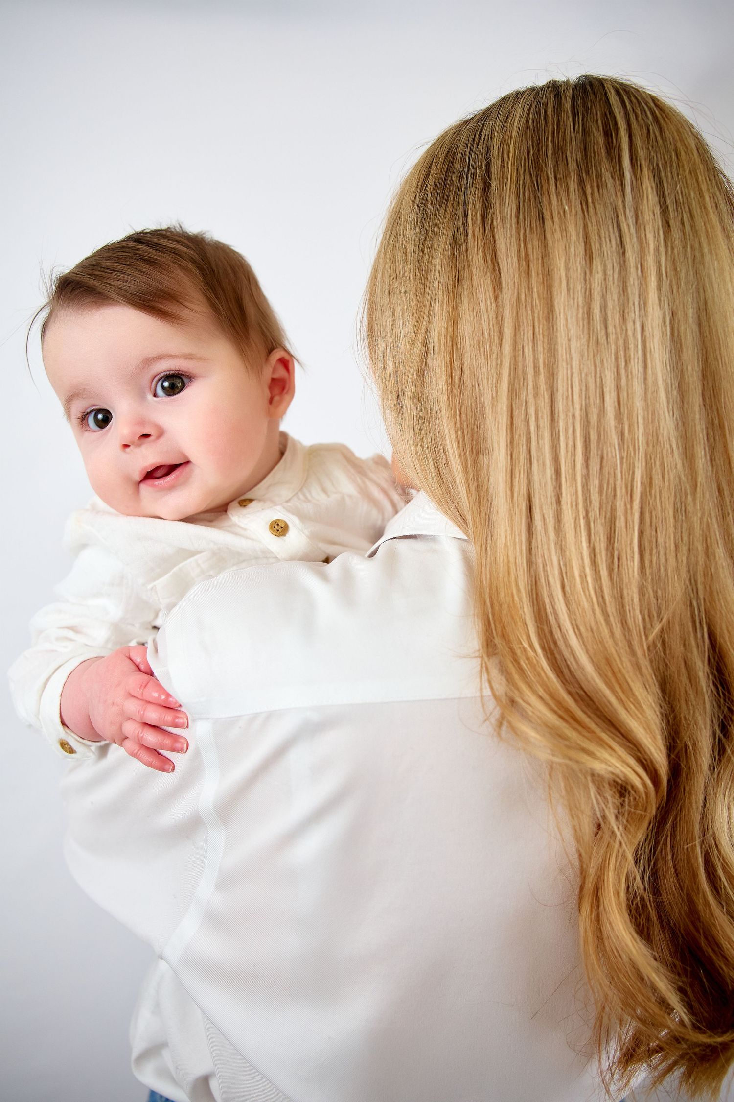A blonde mother holds her smiling baby dressed in white against a light background.