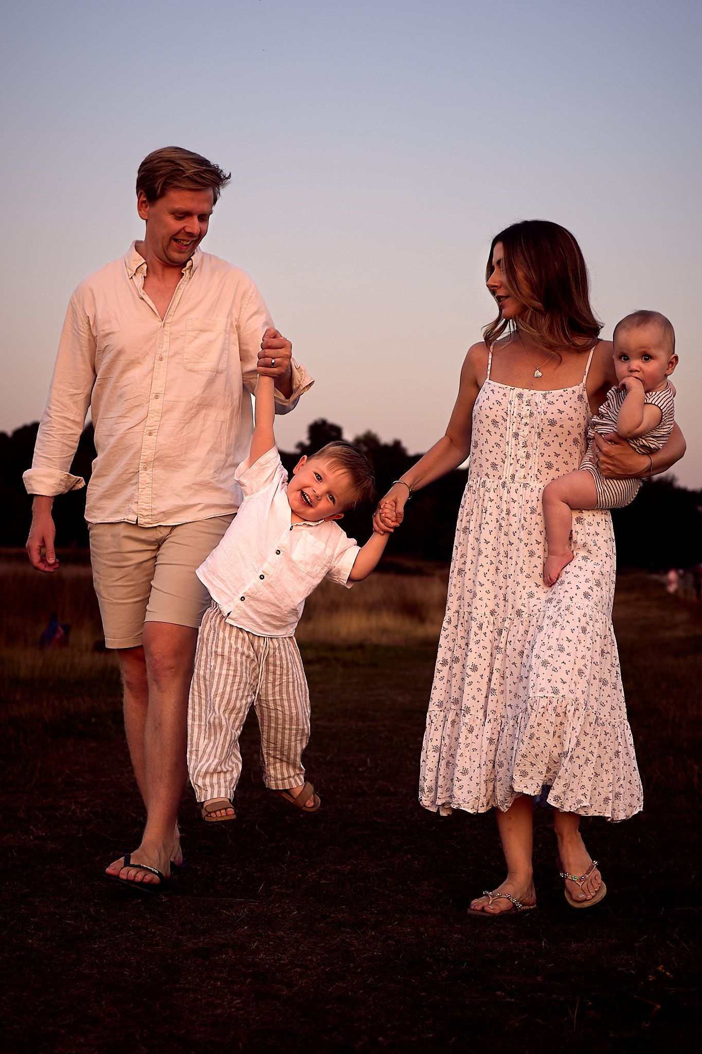 Happy family of four walking in a field at sunset, parents swinging young child between them.
