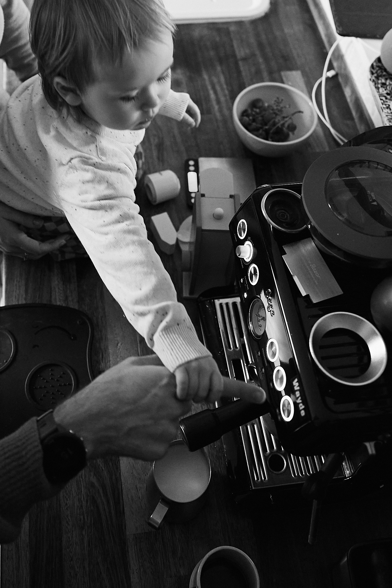 Black and white aerial view of a young child watching an adult operate a home espresso coffee machine.