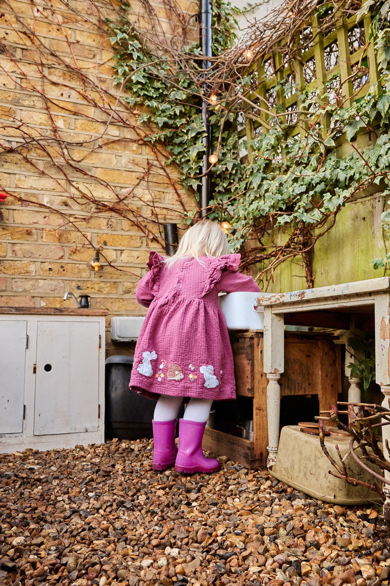 Young girl in pink dress and rain boots playing at an outdoor mud kitchen in a garden.