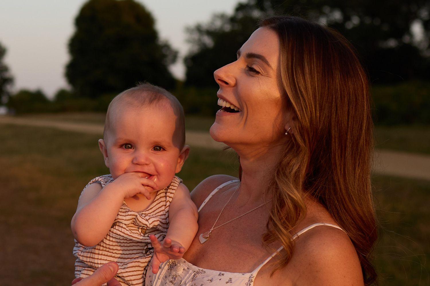 A smiling woman holds a cute baby outdoors at golden hour in a park setting.