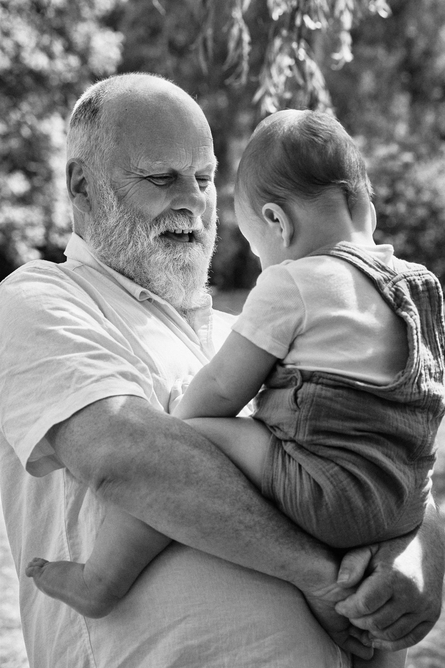 Black and white photo of an elderly bearded man lovingly holding a baby outdoors.