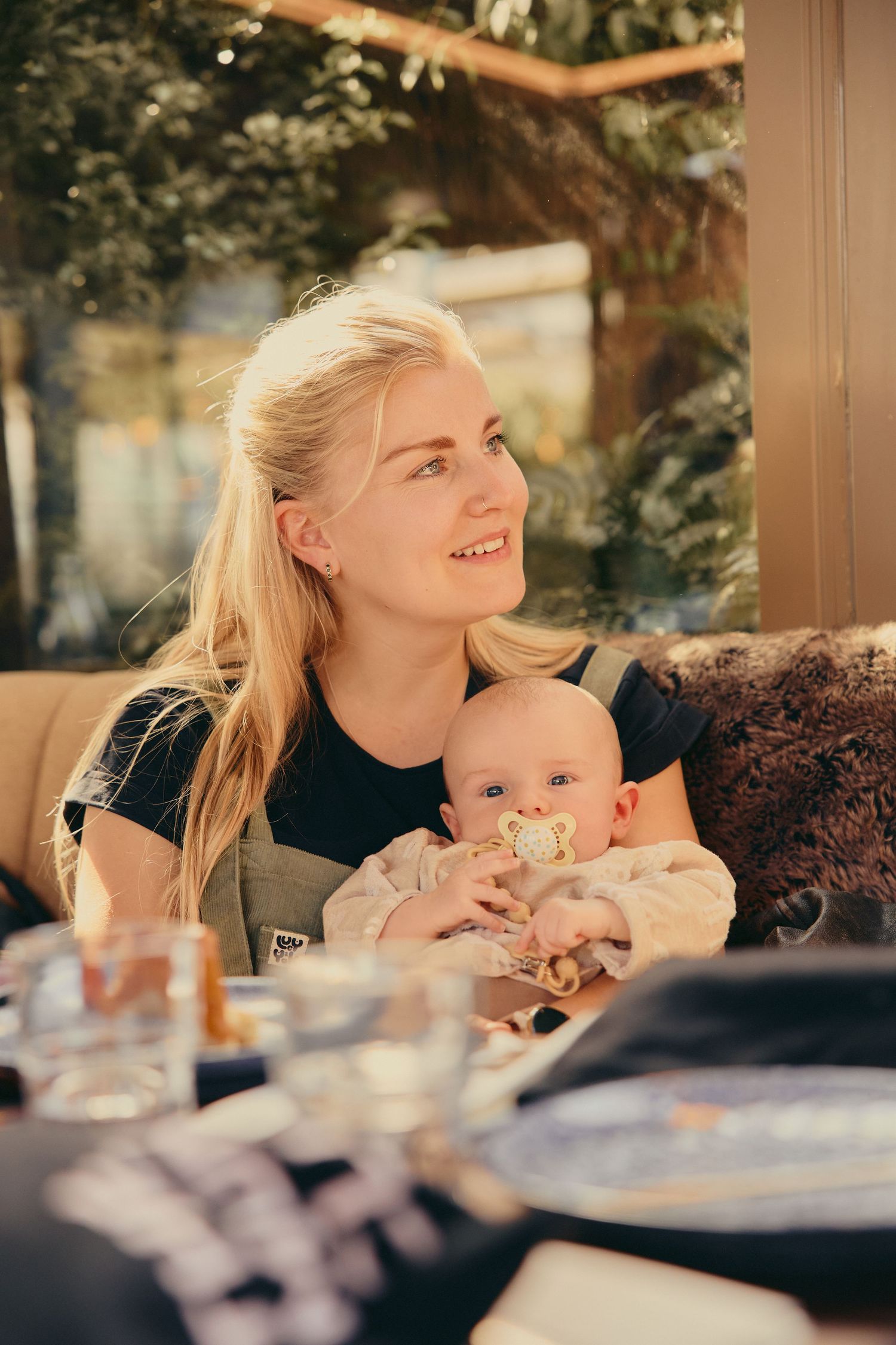 Smiling blonde mother holding her baby with a pacifier at a sunny restaurant table.