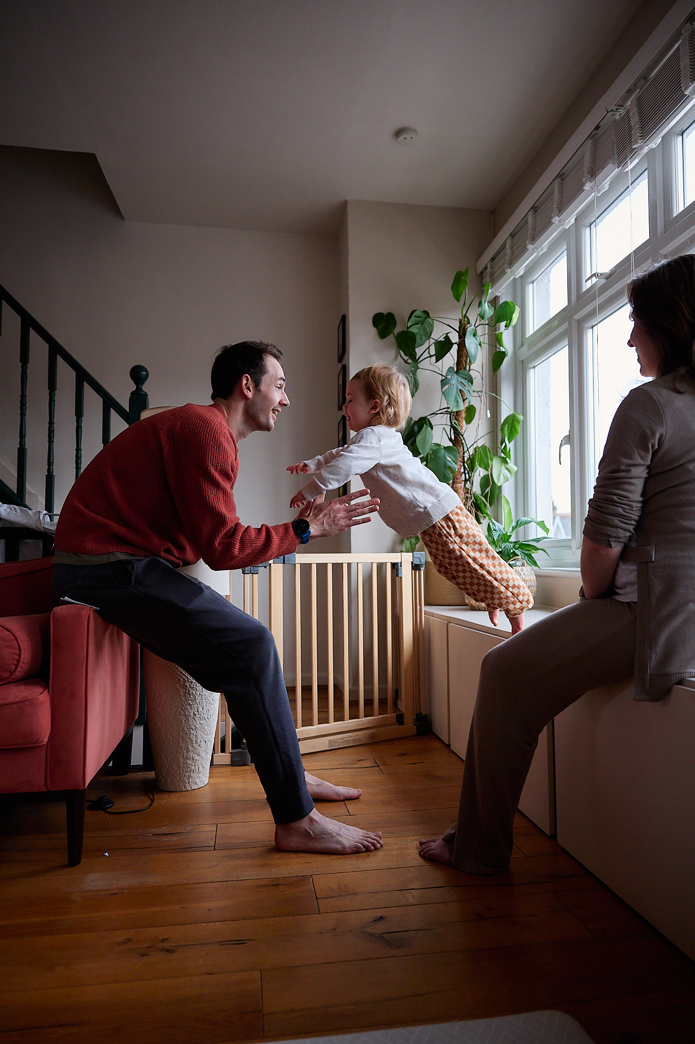 Happy family with toddler playing together in cozy living room, father reaching out to child near window.