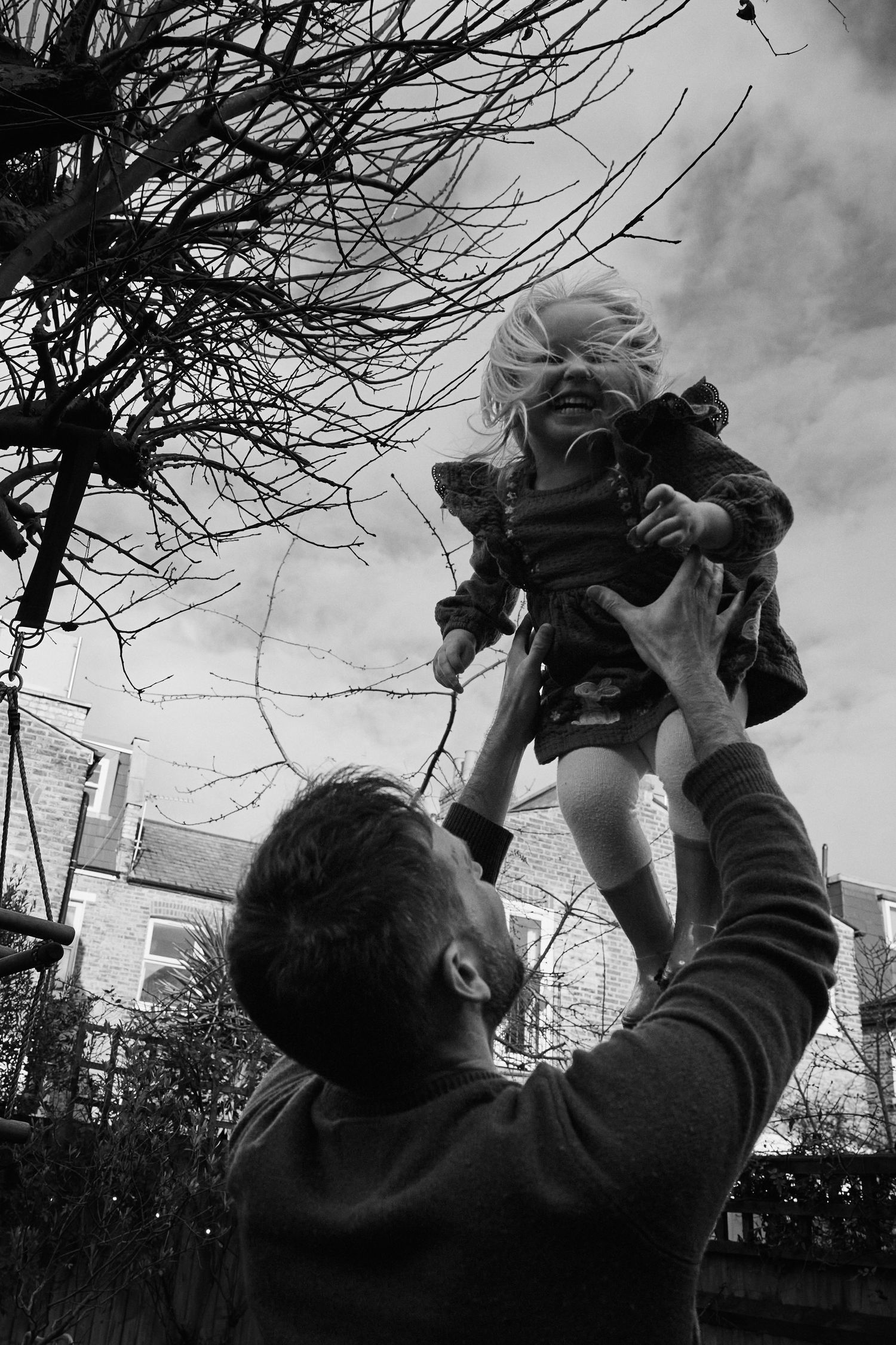 Black and white photo of a father lifting his young blonde child into the air in a garden.