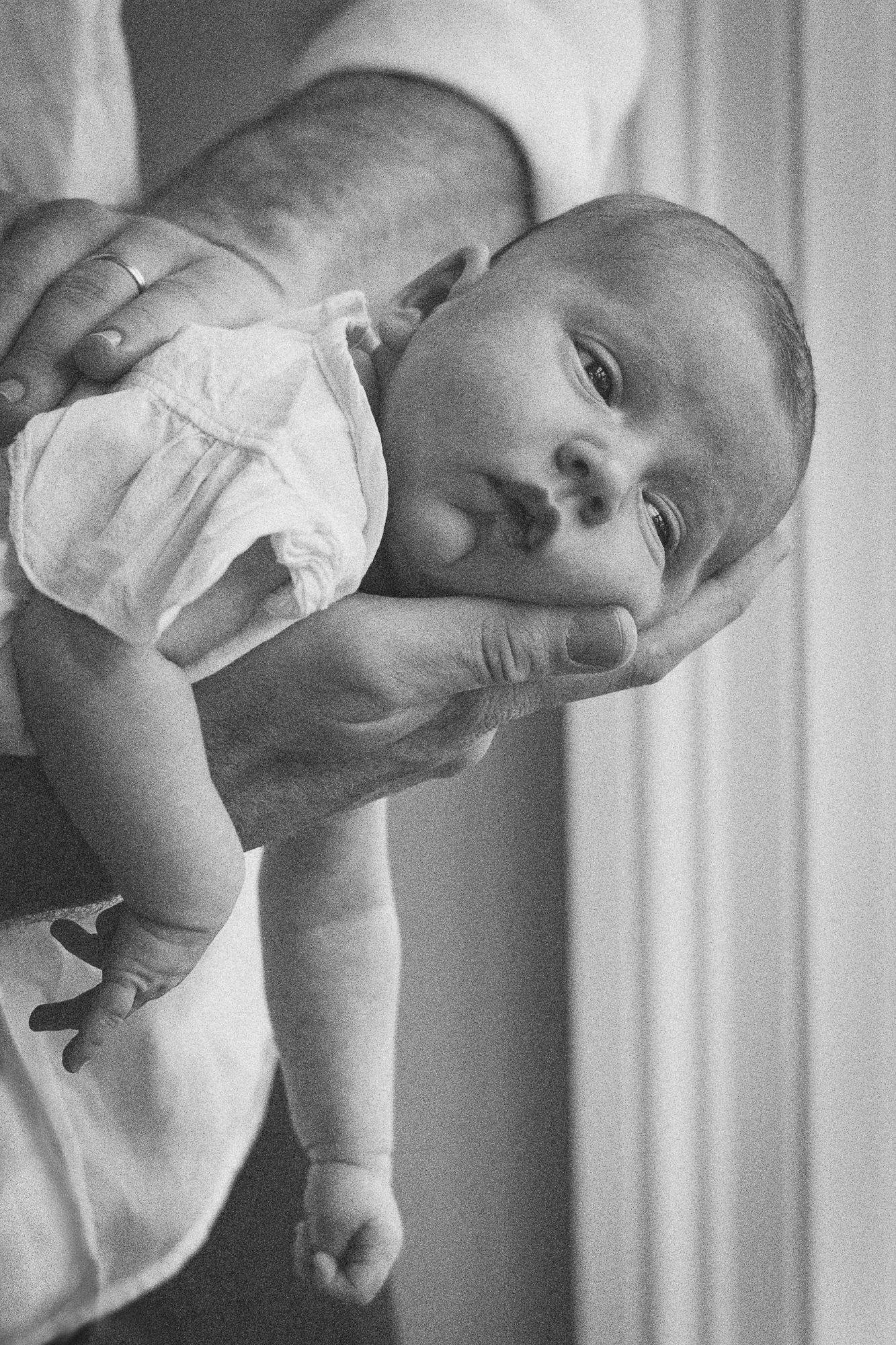 Black and white photo of a newborn baby being held up by an adult's hands, wearing a diaper.
