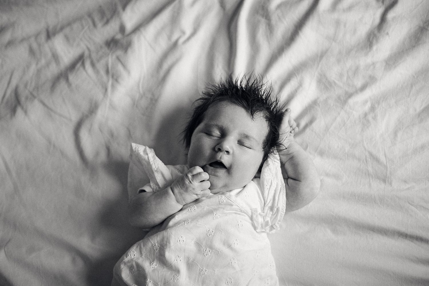 Black and white portrait of an infant with spiky hair sleeping on white bedding.