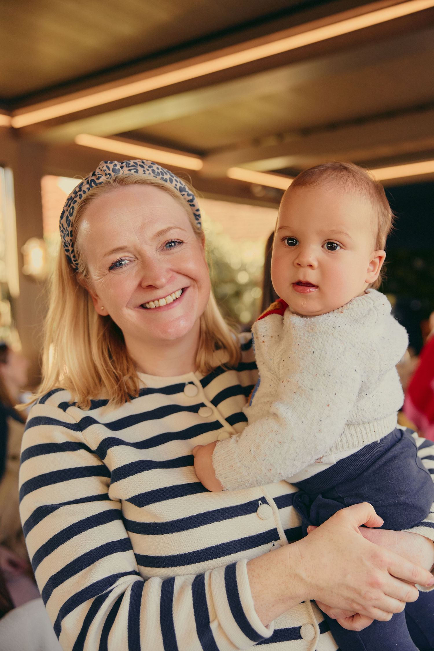 Smiling blonde mother in striped shirt holds her baby at an indoor gathering.