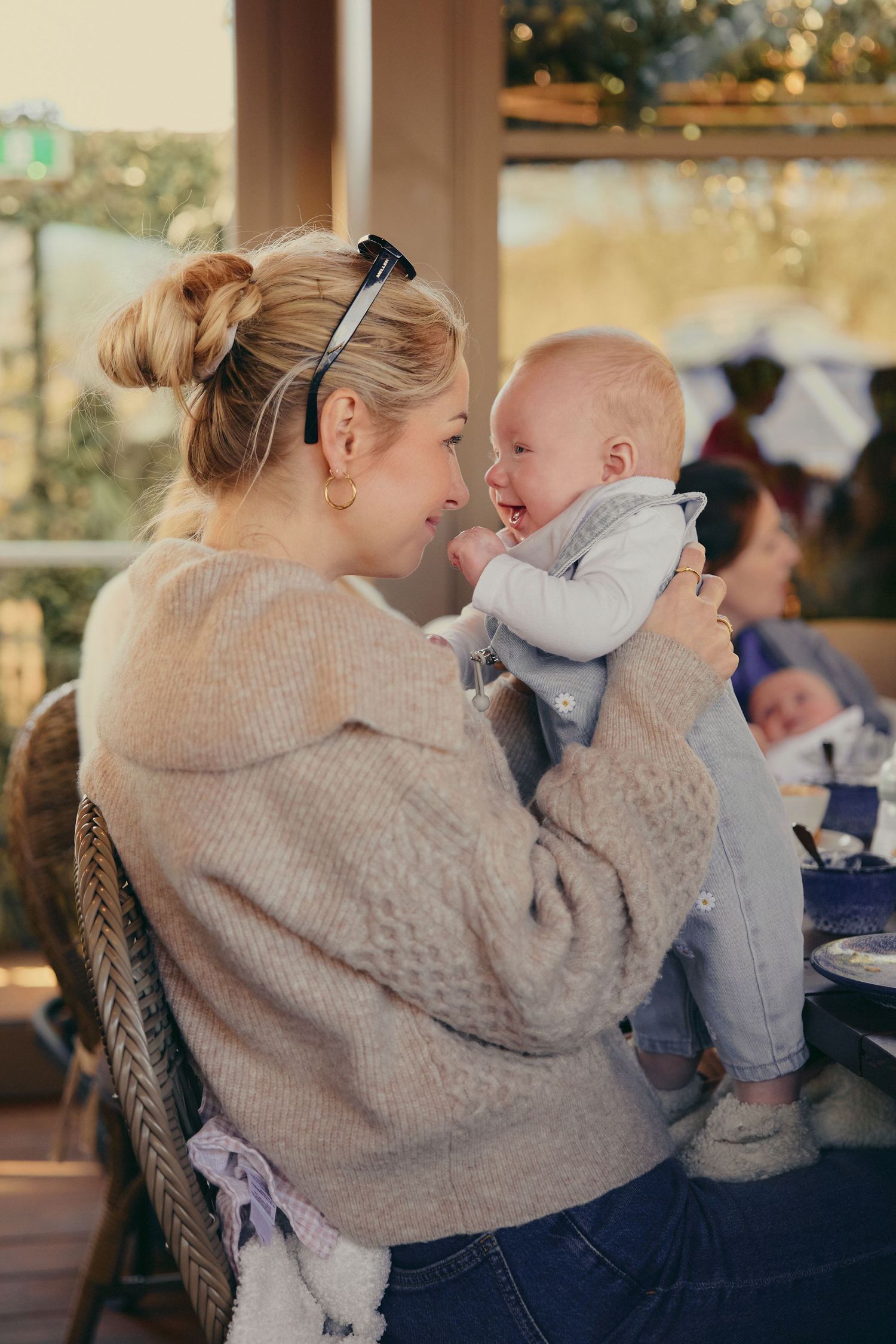 A blonde woman in a beige sweater holds and smiles at a laughing baby on an outdoor patio.