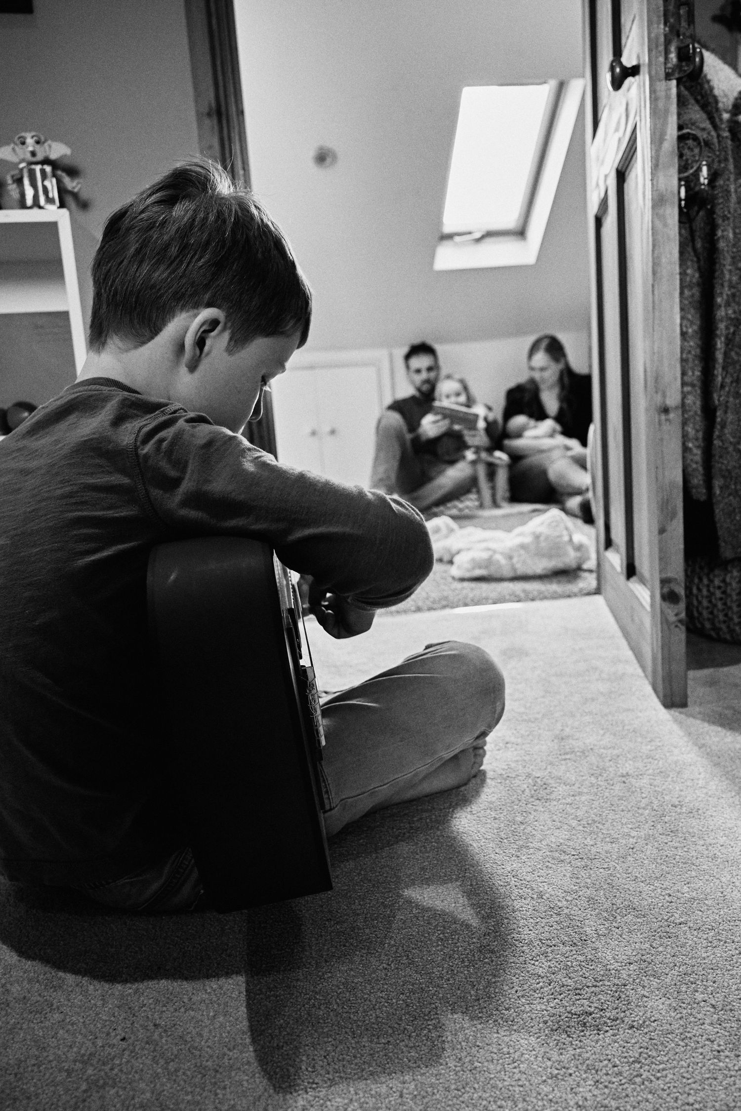 Boy sitting cross-legged on carpet playing guitar in black and white family home photo.