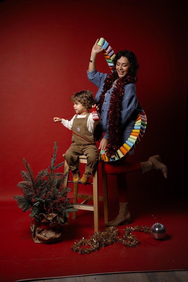 Woman and child posing with Christmas decorations, tinsel, and festive ornaments on a red background.