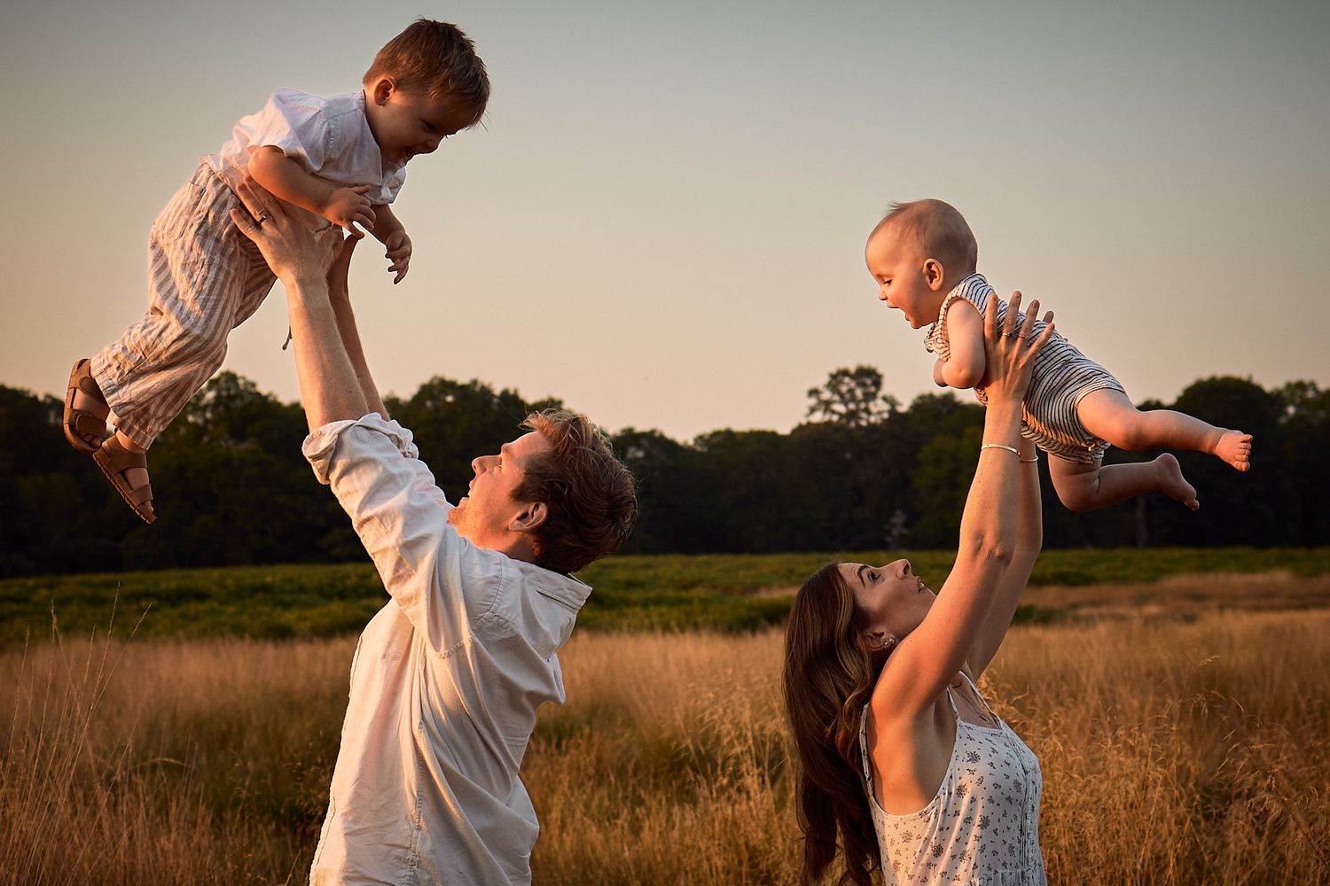 Family playing and lifting child in golden sunset field wearing white and light clothing.