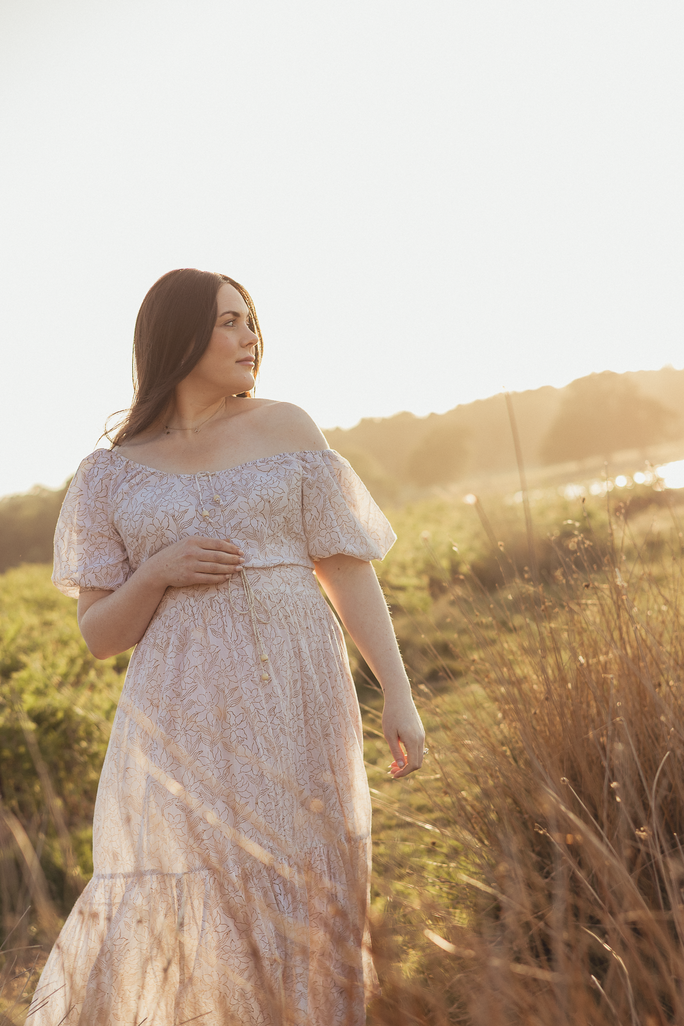 Expectant mother in white lace dress standing in golden sunlit field at sunset.
