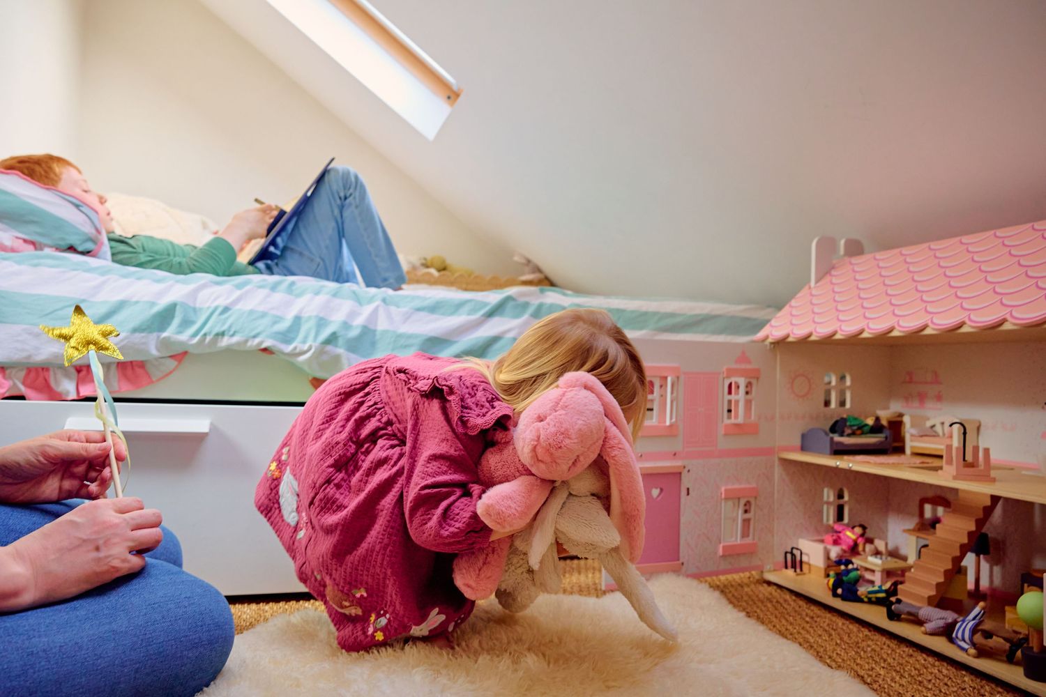 Young girl hugging stuffed bunny beside pink dollhouse in cozy attic bedroom while another child reads on bed.