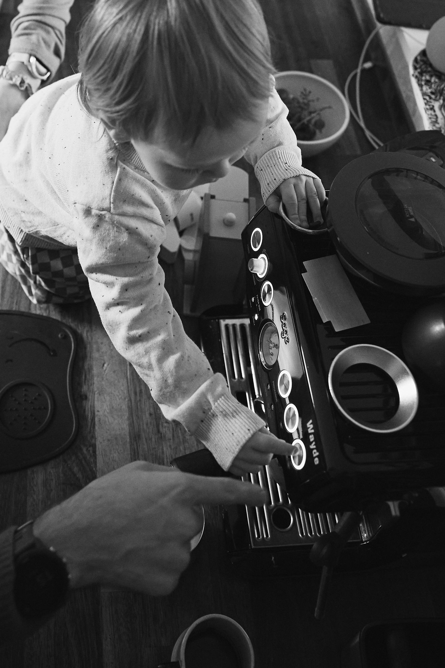 Young child exploring a vintage espresso machine on a wooden table in black and white photo.