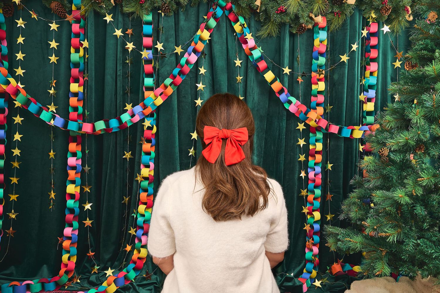 Girl with red bow in hair stands before colorful paper chain garlands and Christmas tree with green backdrop.