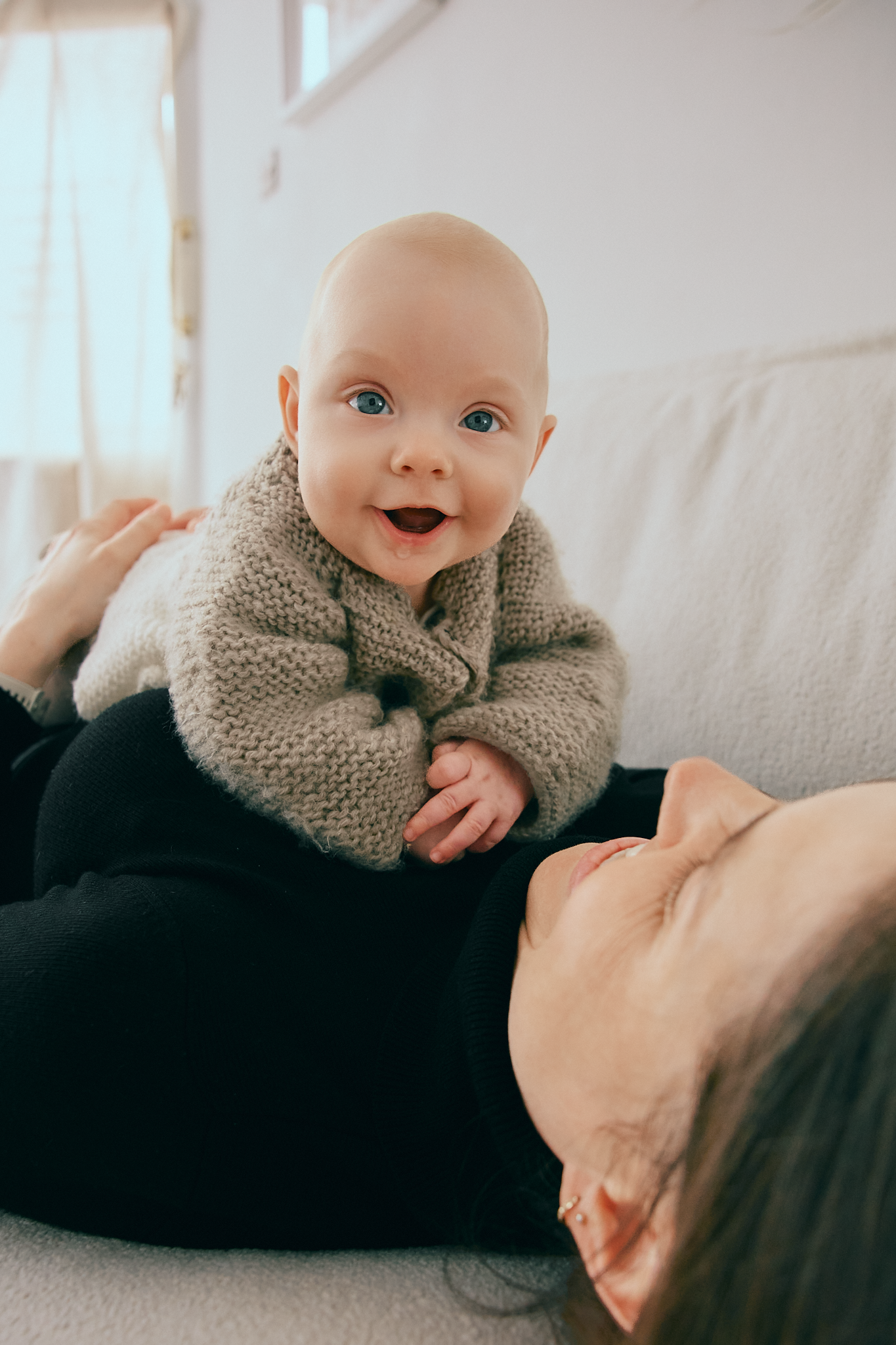 A happy baby in a knit sweater smiles brightly while being held by a mother on a white sofa.