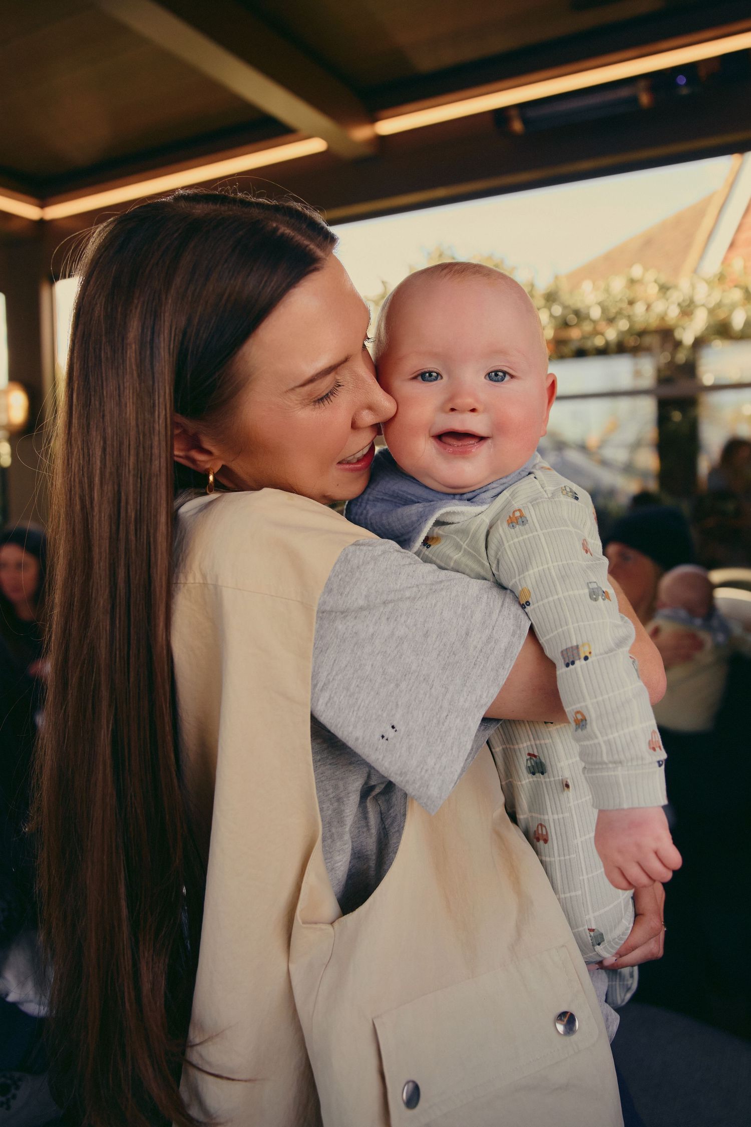 A young mother tenderly kissing her smiling baby boy on the cheek at an indoor venue.