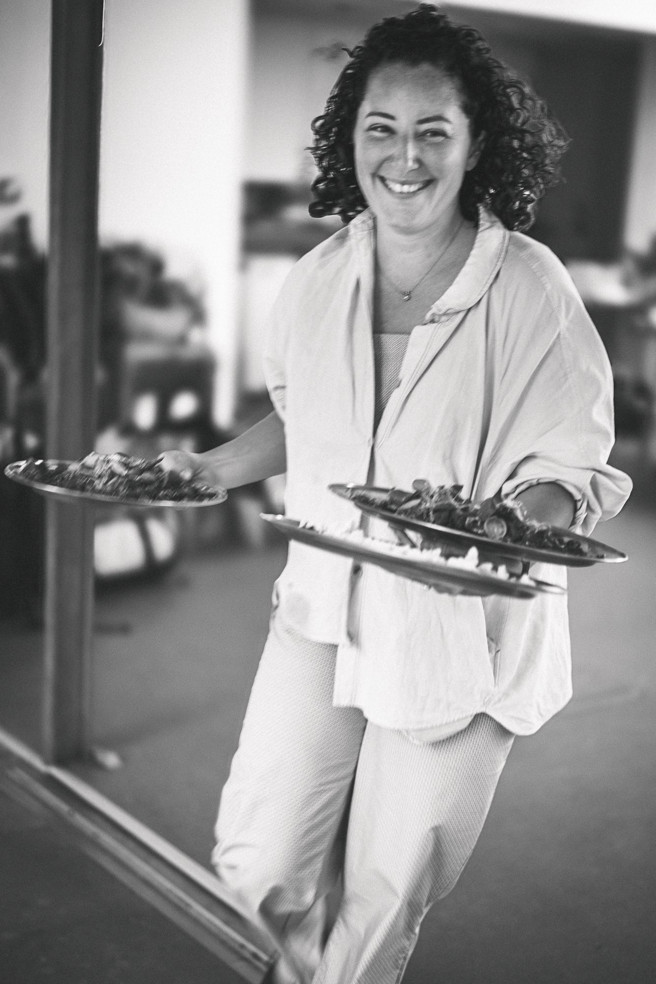 Chef in white attire carries plates of food while smiling in a restaurant kitchen setting.