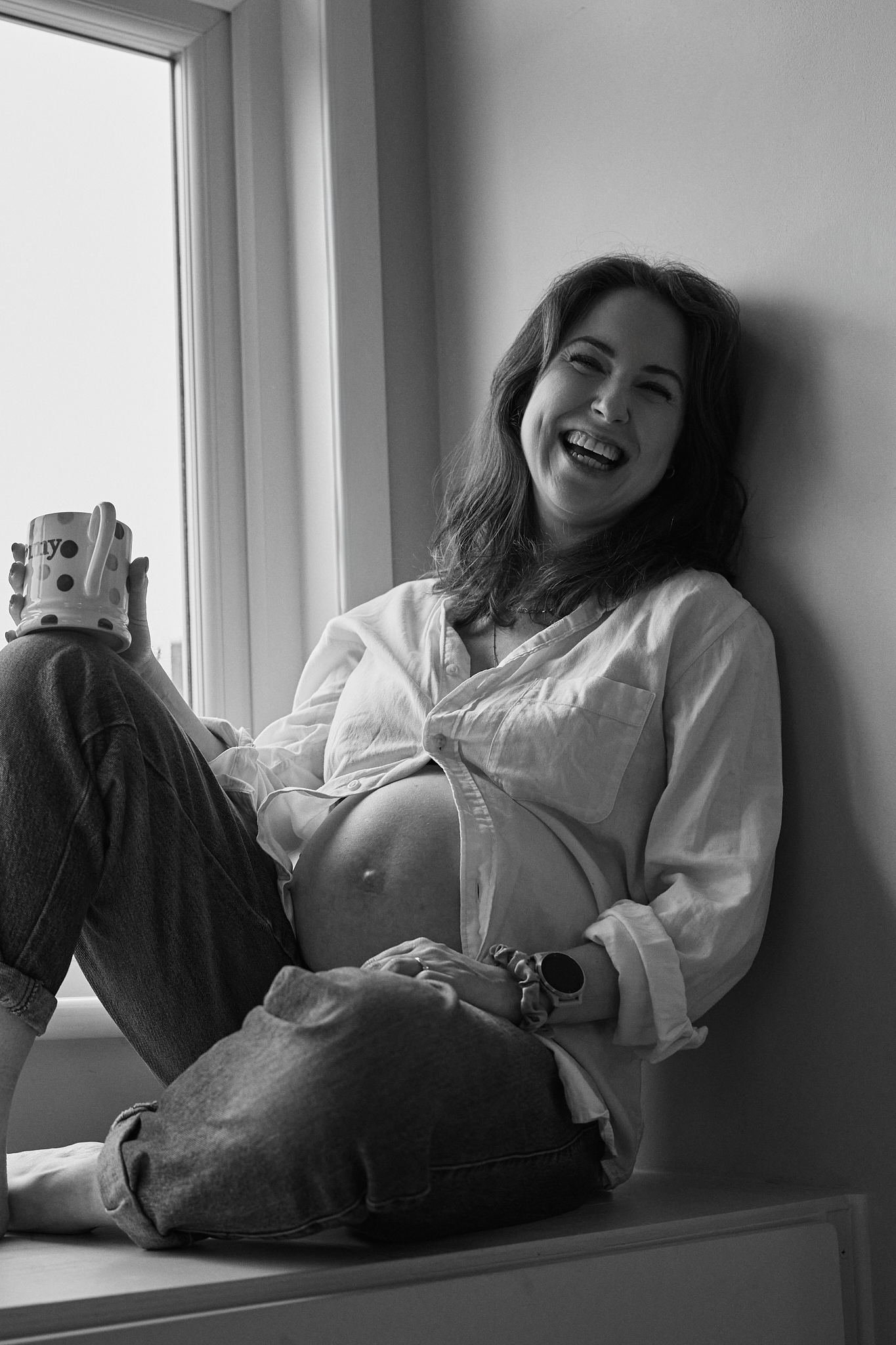 Smiling pregnant woman sitting on windowsill holding a mug, wearing white shirt and jeans, black and white photo.