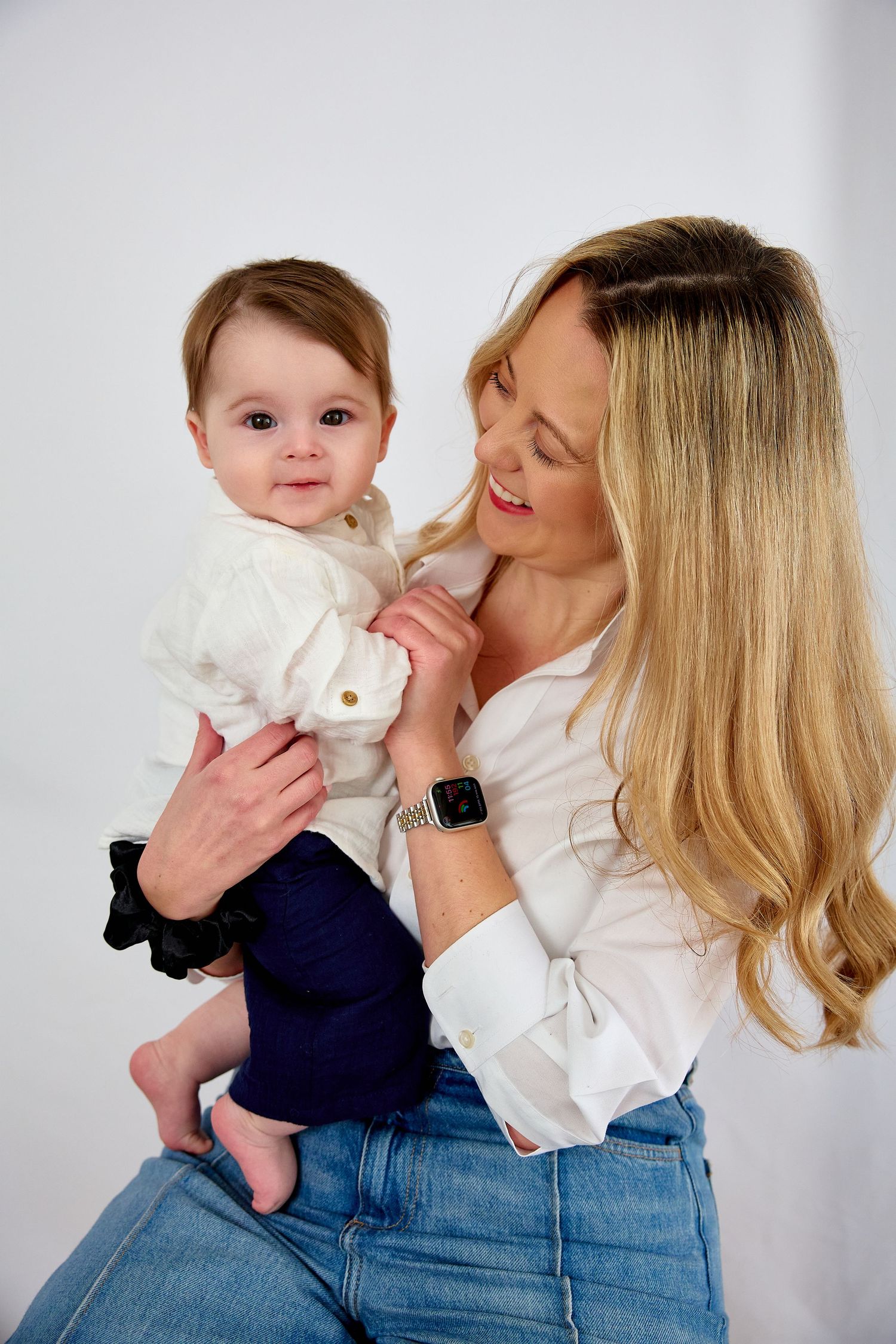 Smiling blonde woman in white shirt holding a cute baby in white top and navy pants against white background.