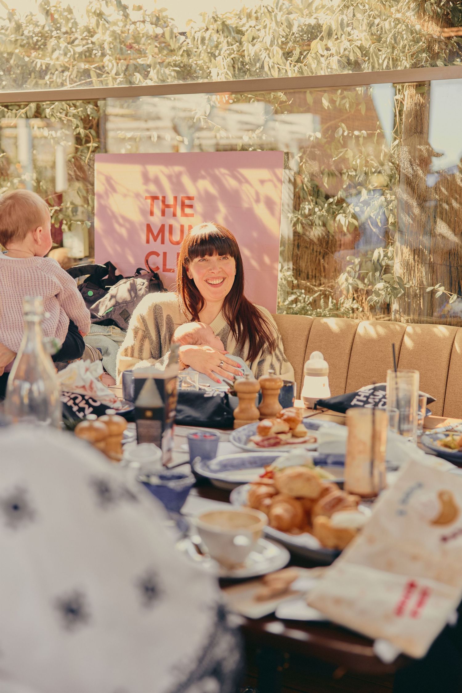 Smiling woman sitting at a table with food and drinks at The Mum Club event in a sunny cafe setting.
