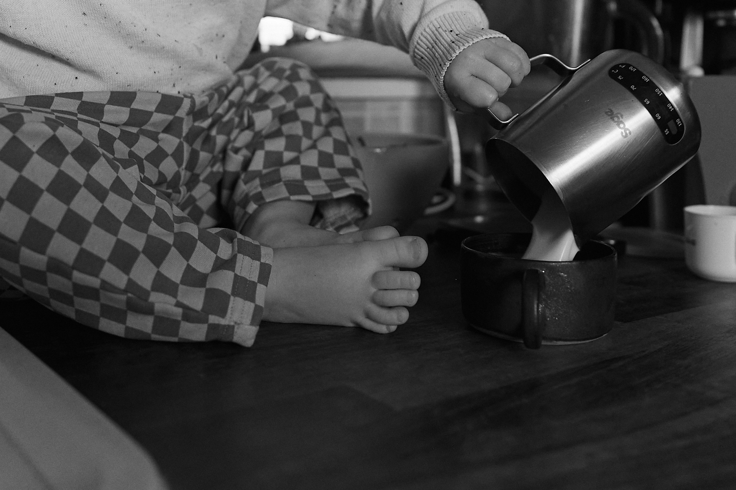 Black and white photo of a child in checkered pants pouring liquid from a small pitcher on a kitchen counter.