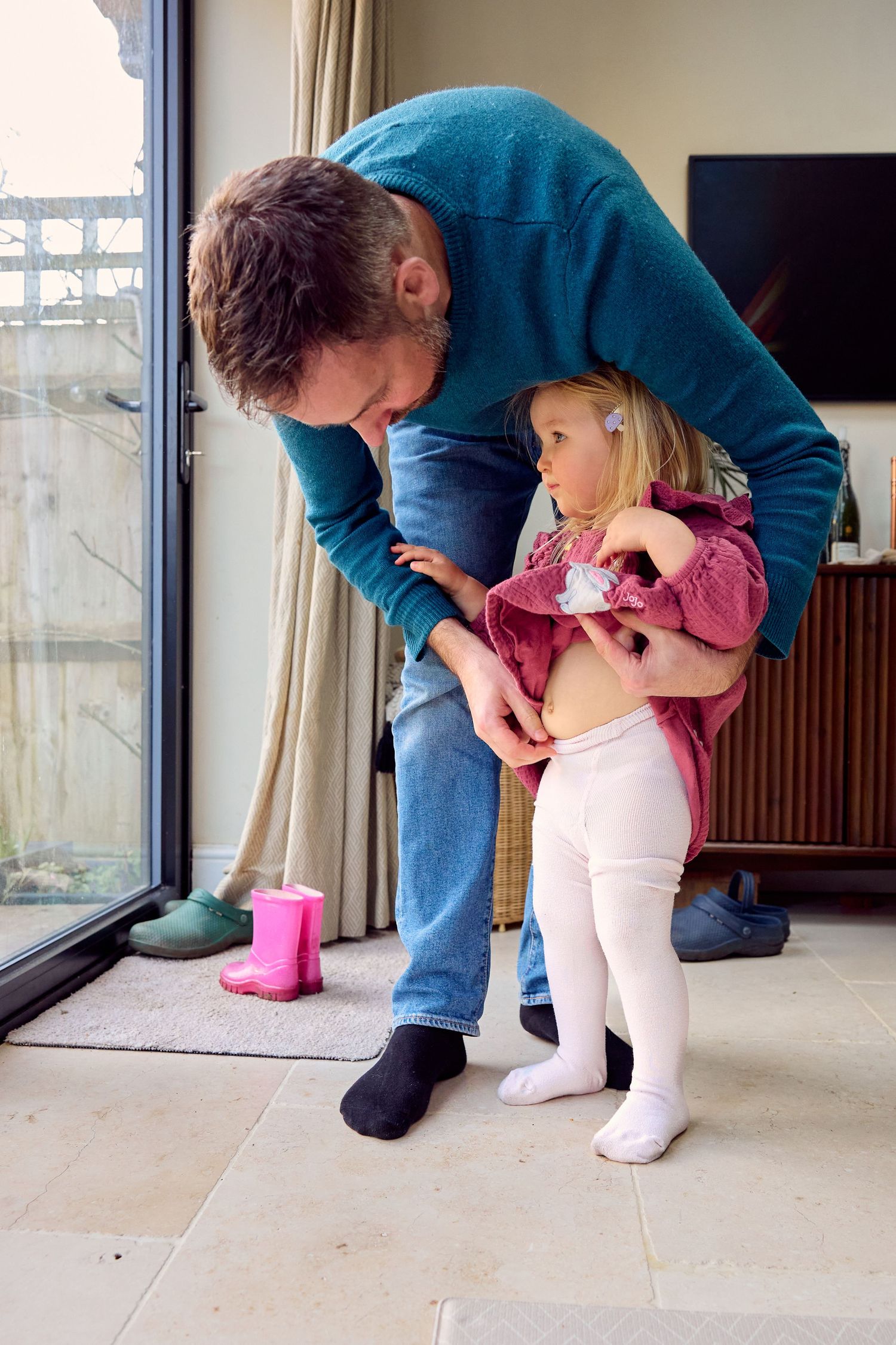 Father helping young toddler girl get dressed near sliding glass door at home.