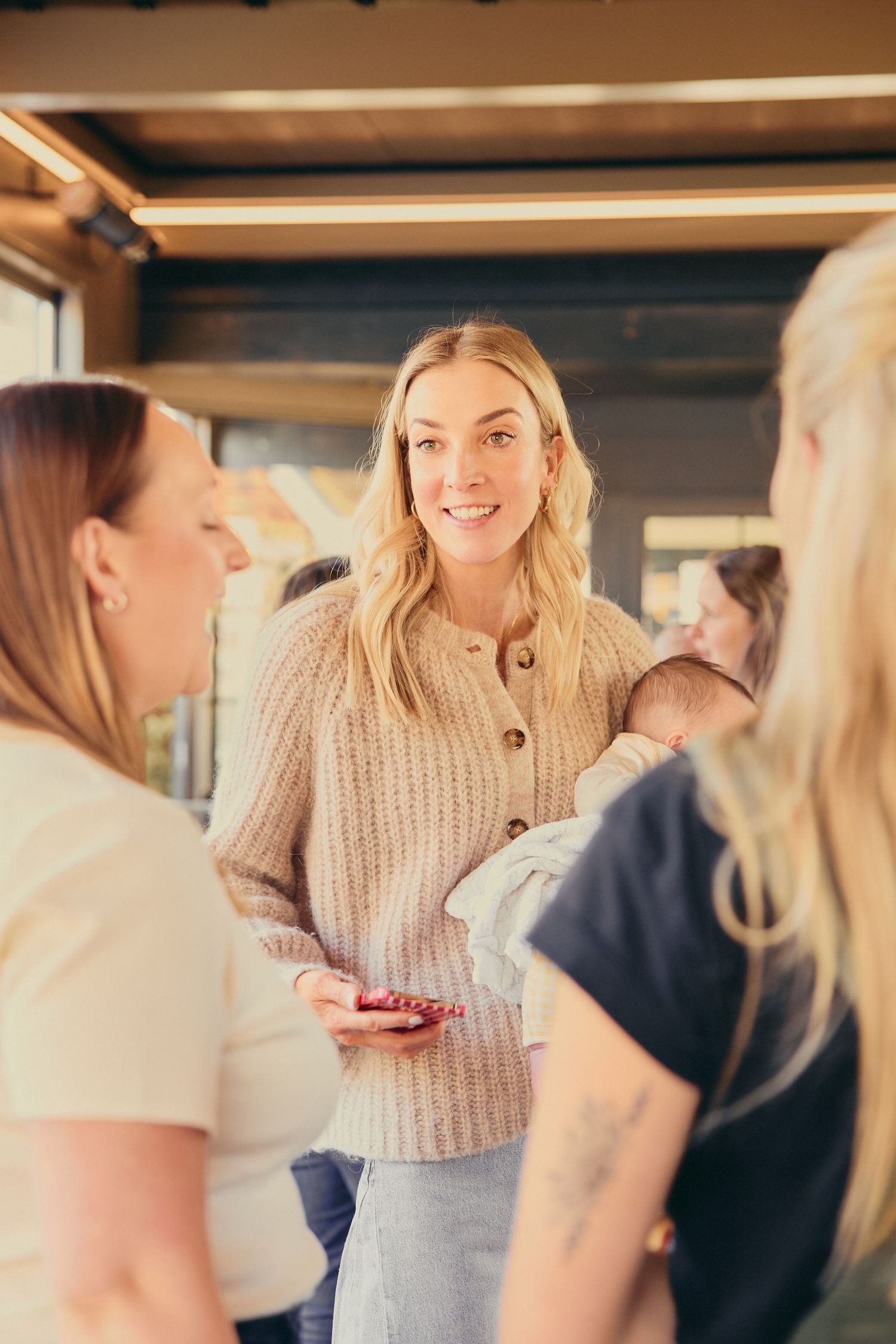 Blonde woman in beige knit cardigan socializes at an indoor gathering with friends.