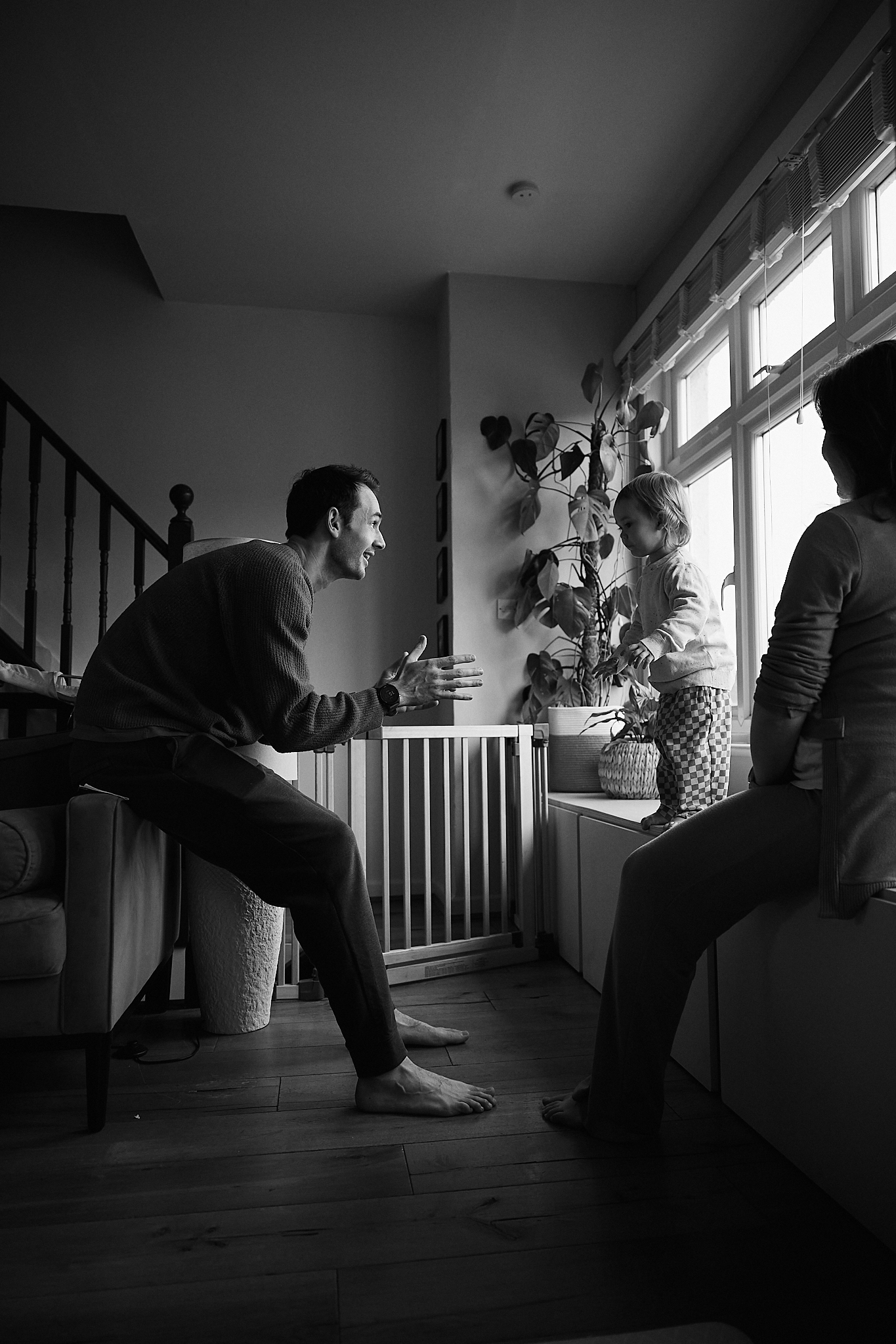 Black and white photo of a father playfully interacting with his children in a cozy home living room near a staircase.